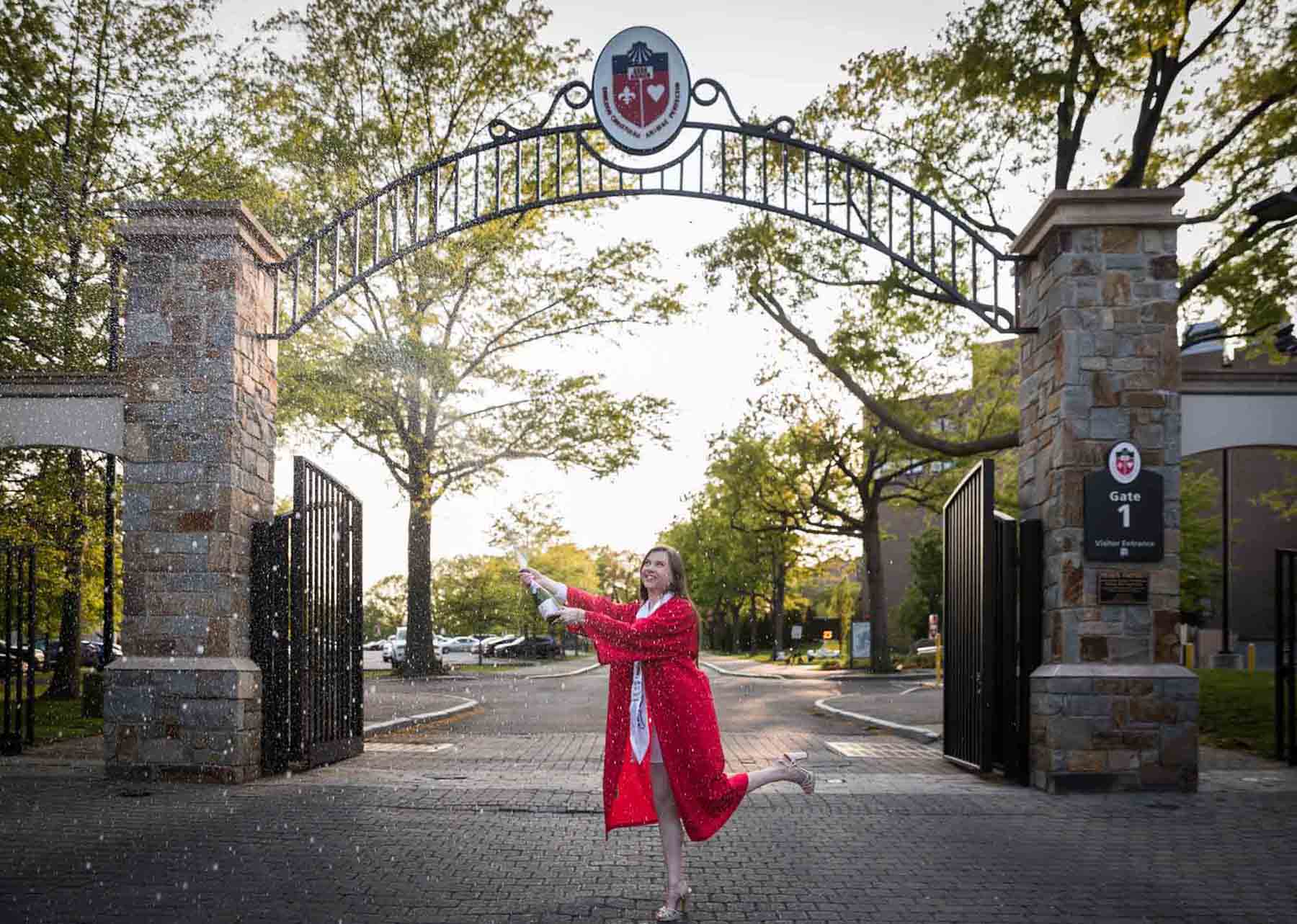 Female graduate wearing red robe and white sash spraying champagne bottle under gate during a St. John’s University graduate portrait session