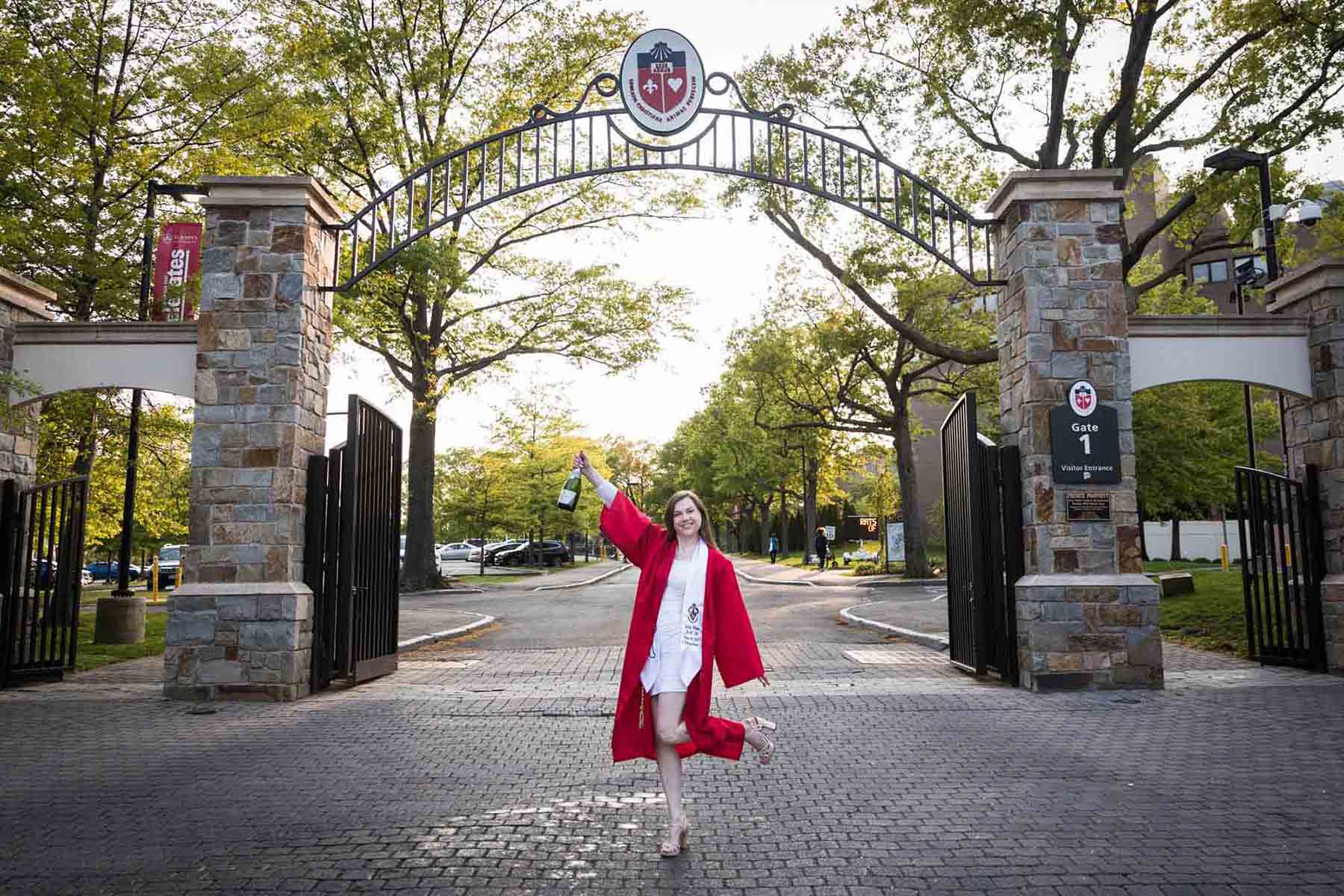 Female graduate wearing red robe and white sash holding champagne bottle under gate during a St. John’s University graduate portrait session