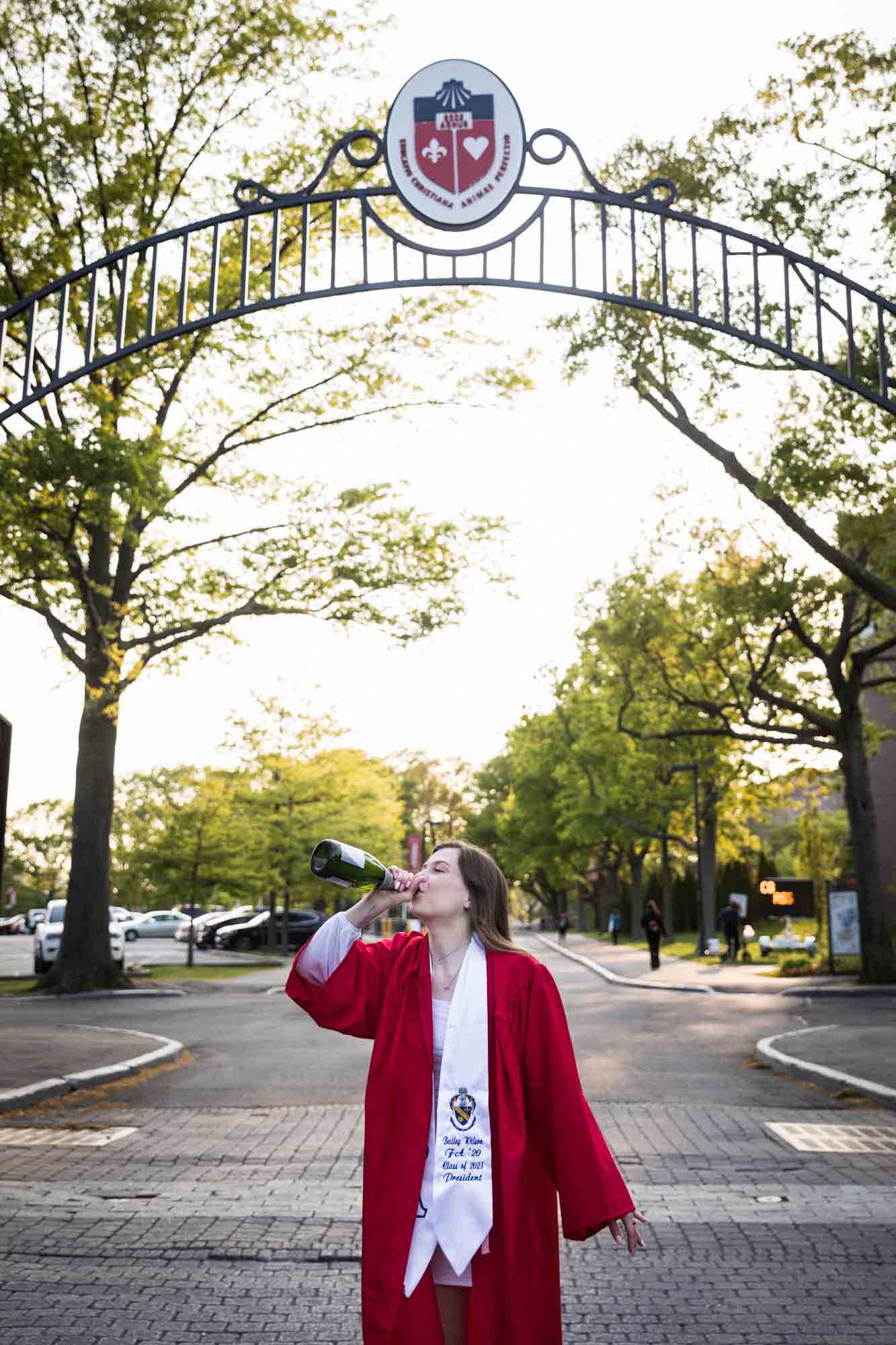 Female graduate wearing red robe and white sash drinking from champagne bottle under gate during a St. John’s University graduate portrait session