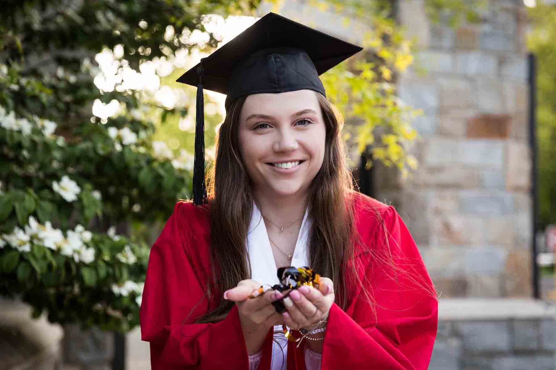 Female graduate wearing red robe and cap holding confetti into the camera for an article on graduation portrait photo tips