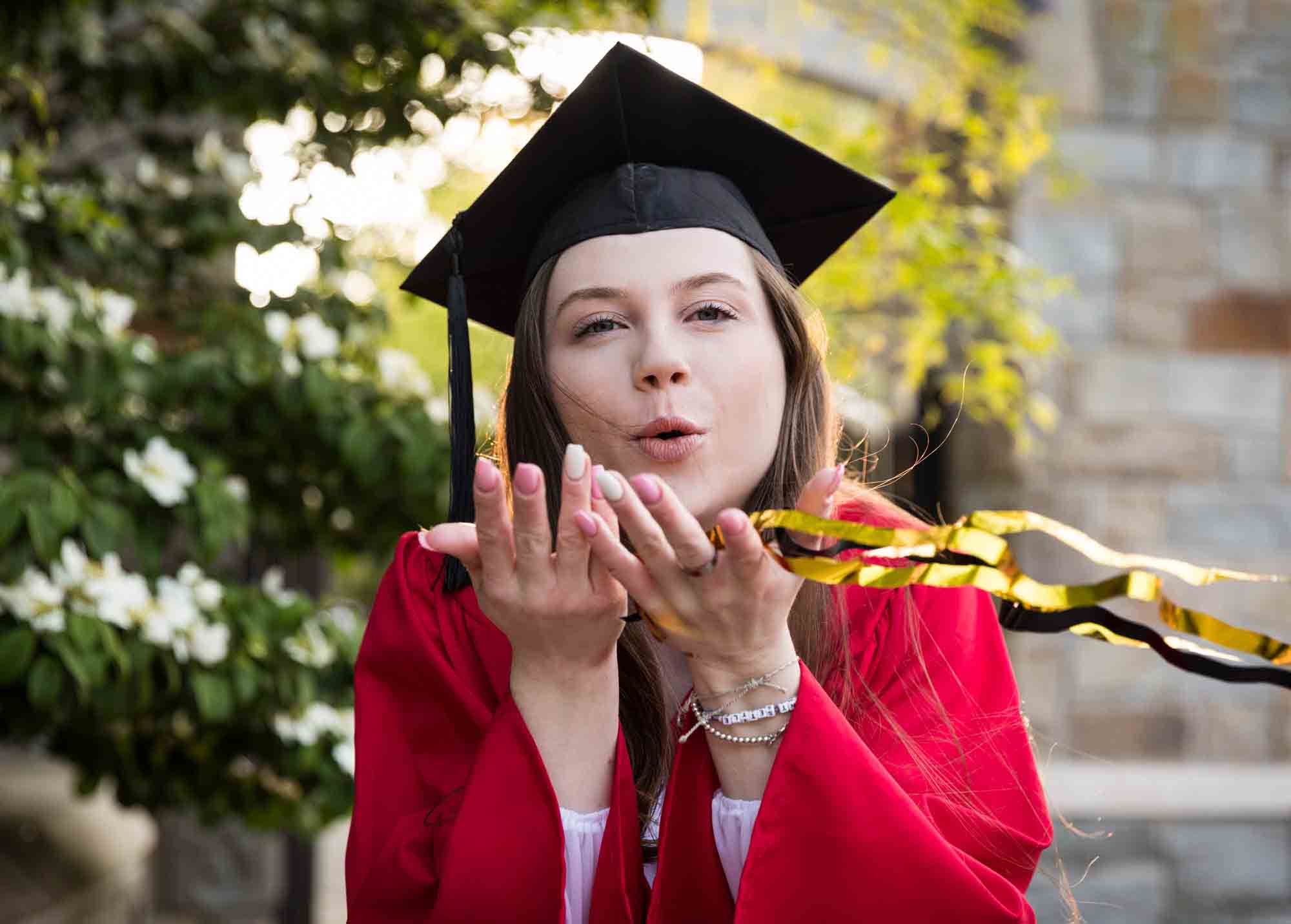 Female graduate wearing red robe and cap blowing confetti into the camera for an article on graduation portrait photo tips