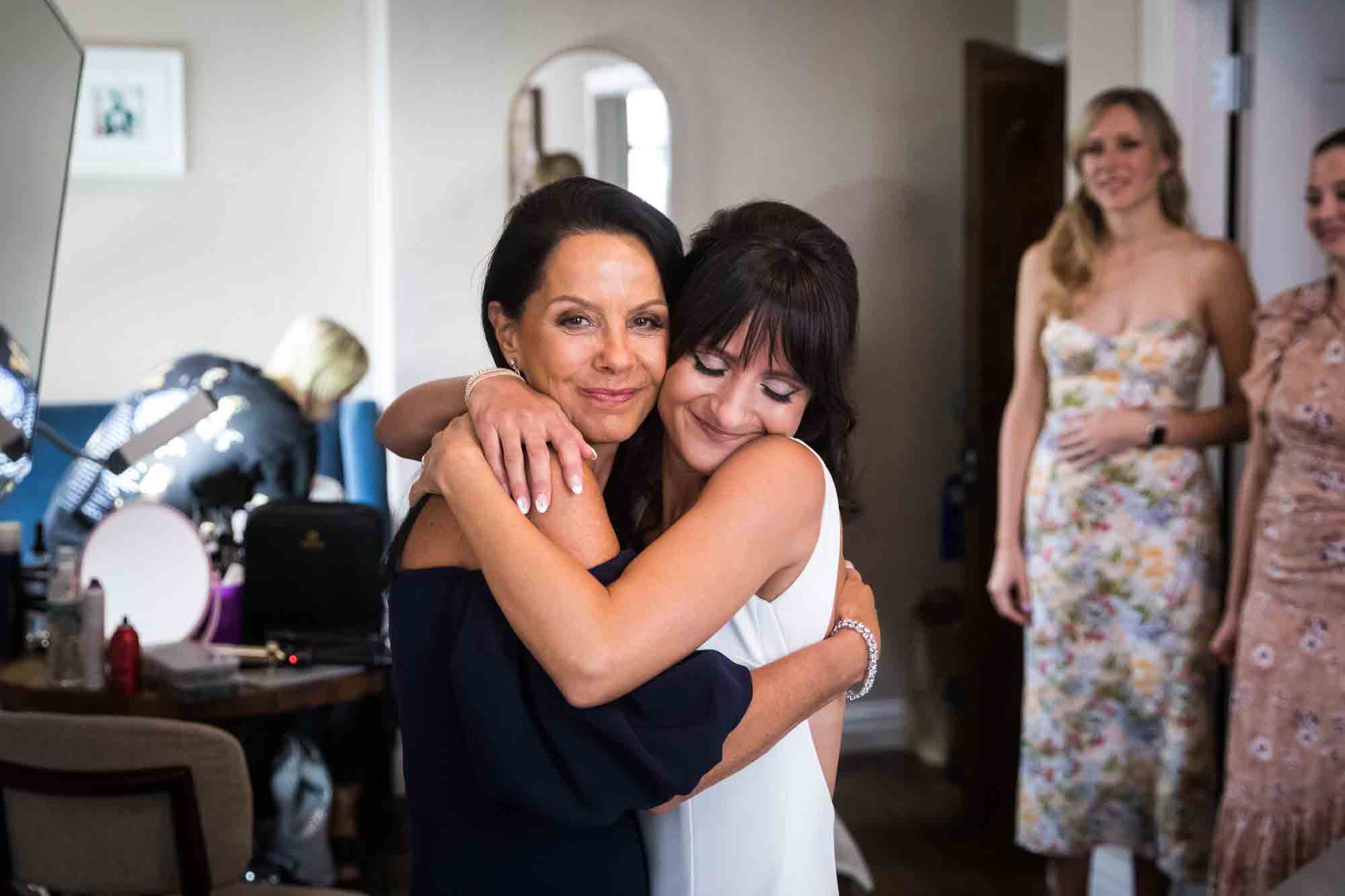 Mother hugging bride in front of two bridesmaids in hotel room