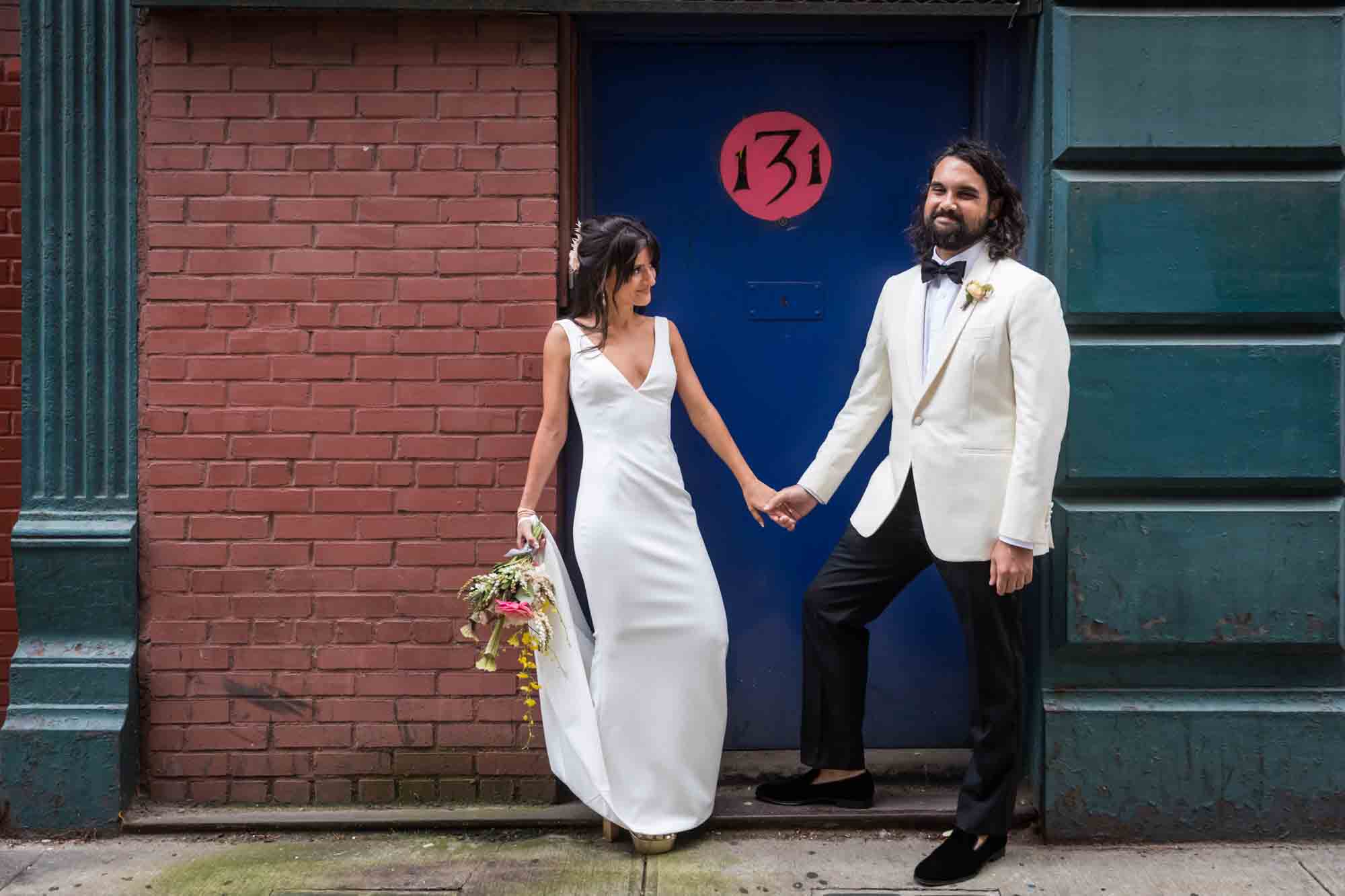 Bride and groom holding hands in front of blue door with number 131 in red circle