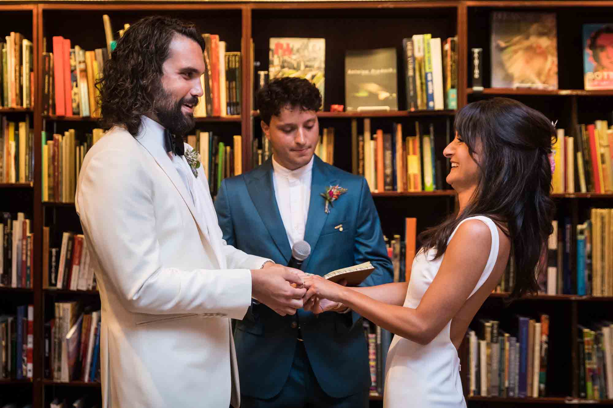 Groom putting ring on bride's finger in front of bookcase at a Housing Works wedding