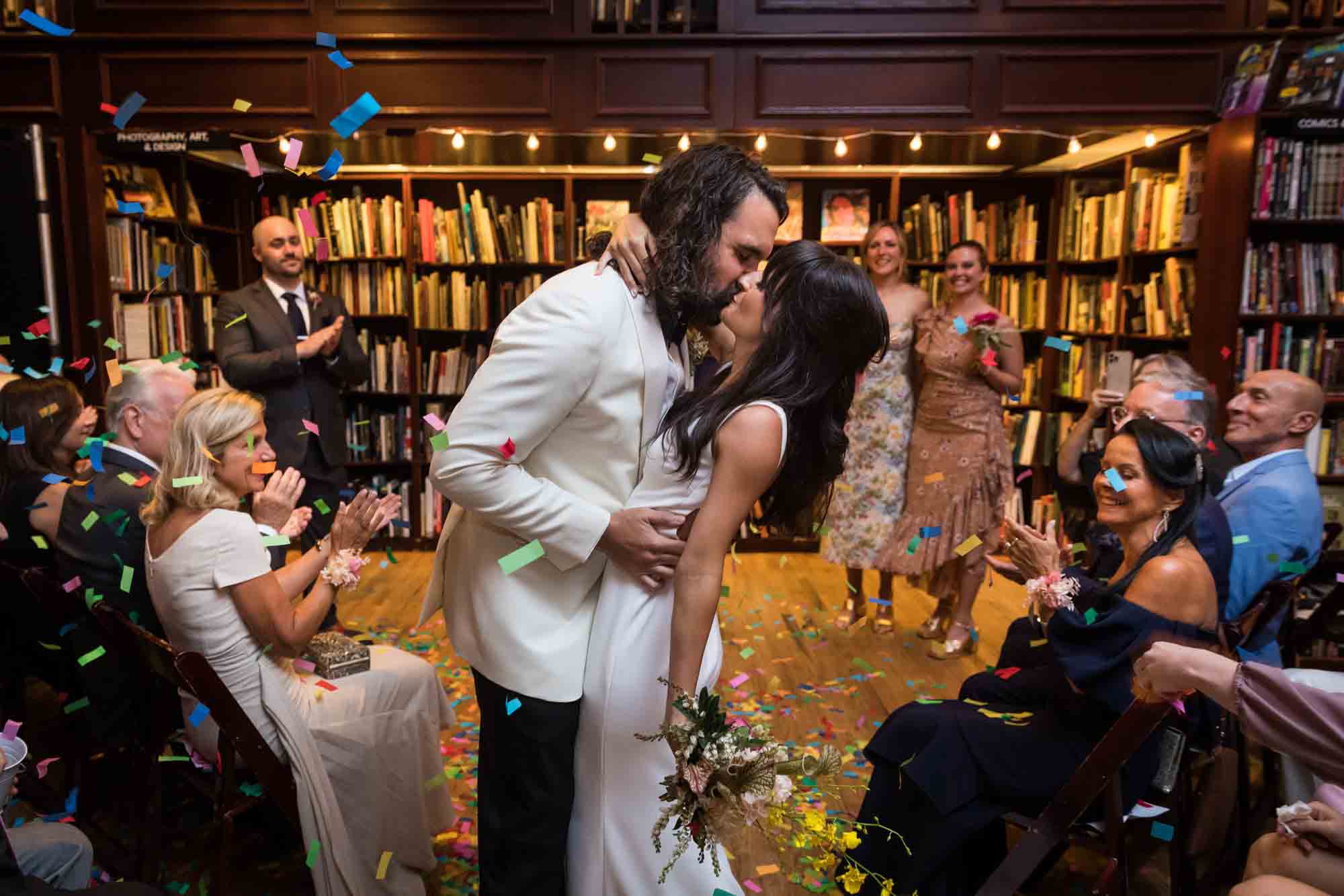 Bride and groom kissing in aisle as guests clap and throw confetti at a Housing Works wedding