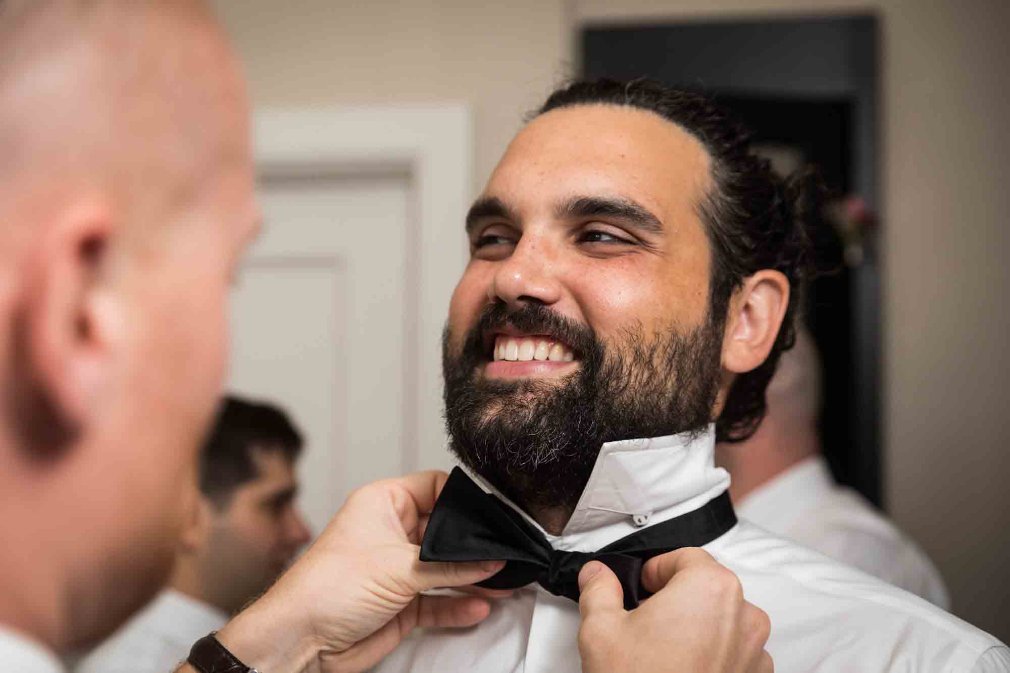 Groomsman helping smiling groom tie bow tie in hotel room