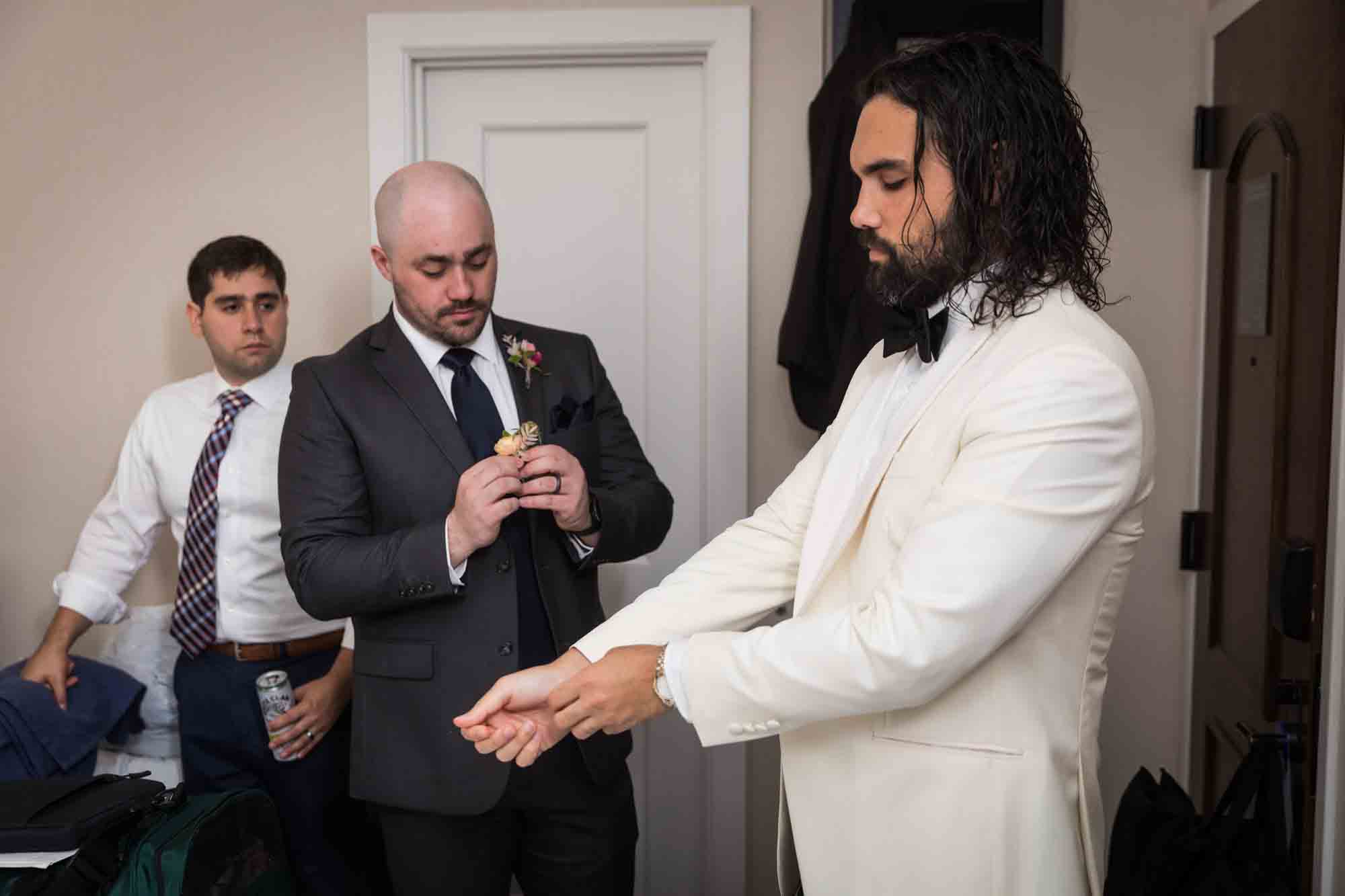 Groom pulling on sleeve of white jacket in front of groomsman holding flowers in hotel room