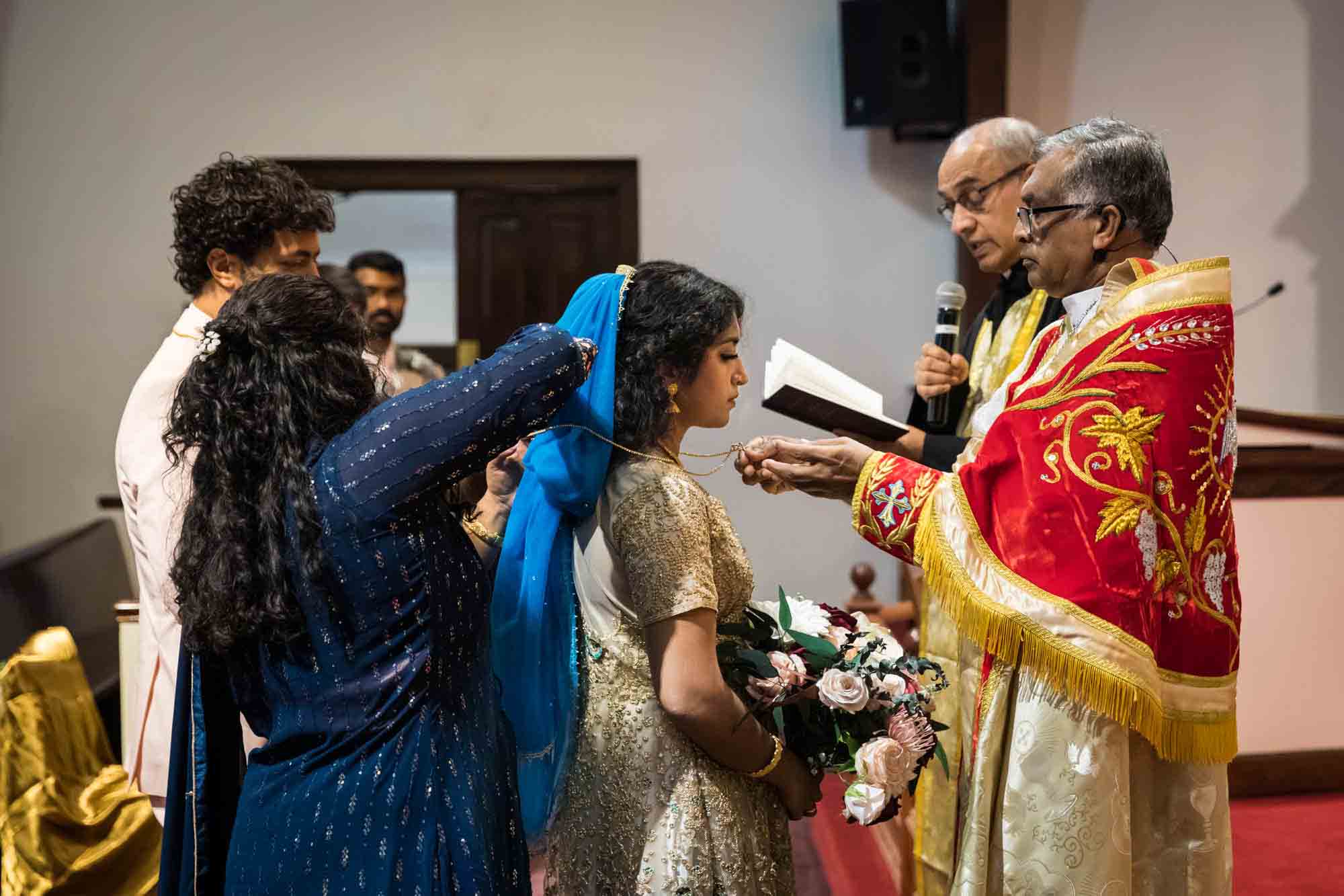 Bridesmaid helping to put gold necklace around bride during Syro-Malankara Catholic wedding ceremony