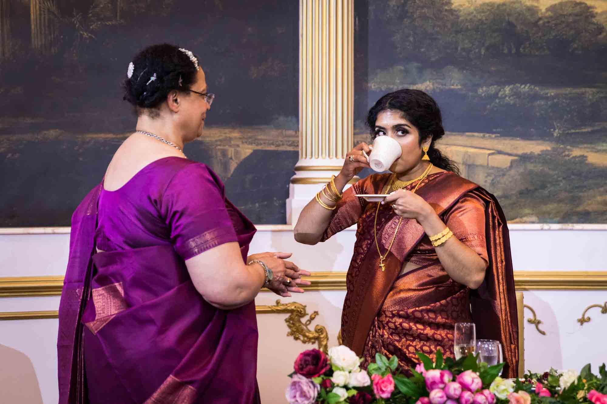 Indian bride wearing rust colored sari and drinking from teacup in front of older woman wearing pink sari during a Terrace on the Park wedding