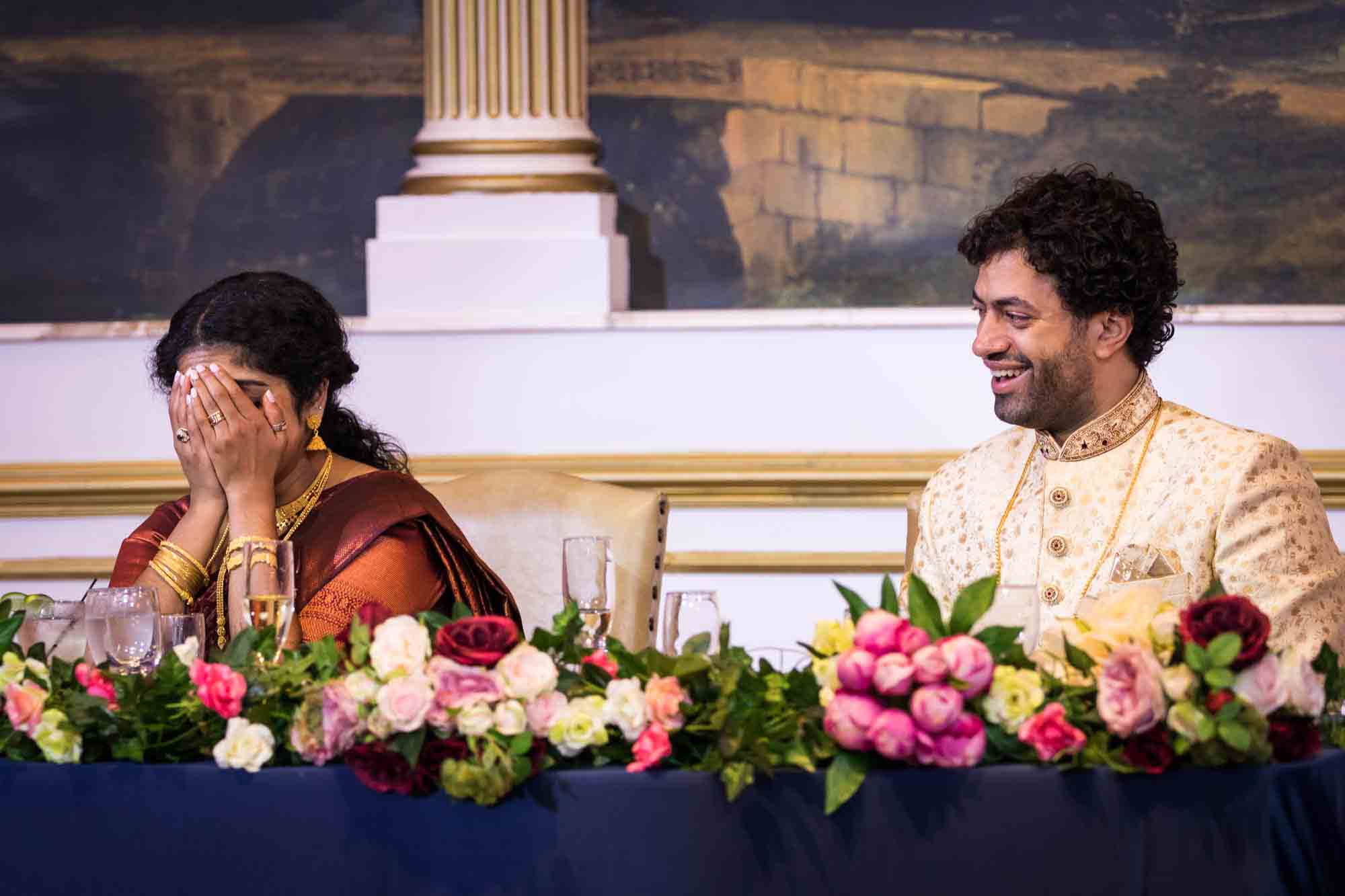 Groom sitting at table watching bride with hands over her face during speeches for an article on Terrace on the Park wedding photo tips