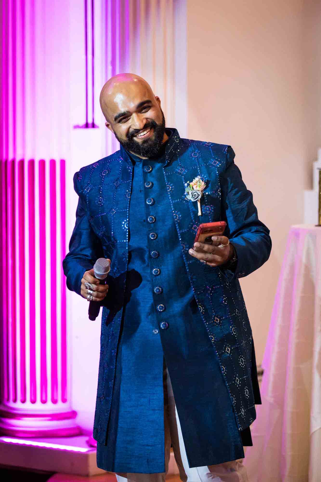 Bald groomsman wearing blue tunic holding microphone and cell phone during speech for an article on Terrace on the Park wedding photo tips
