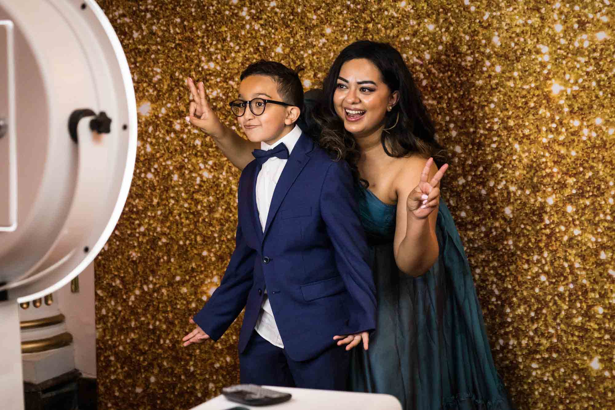 Woman standing behind boy wearing bow tie in front of gold background of photo booth for an article on Terrace on the Park wedding photo tips
