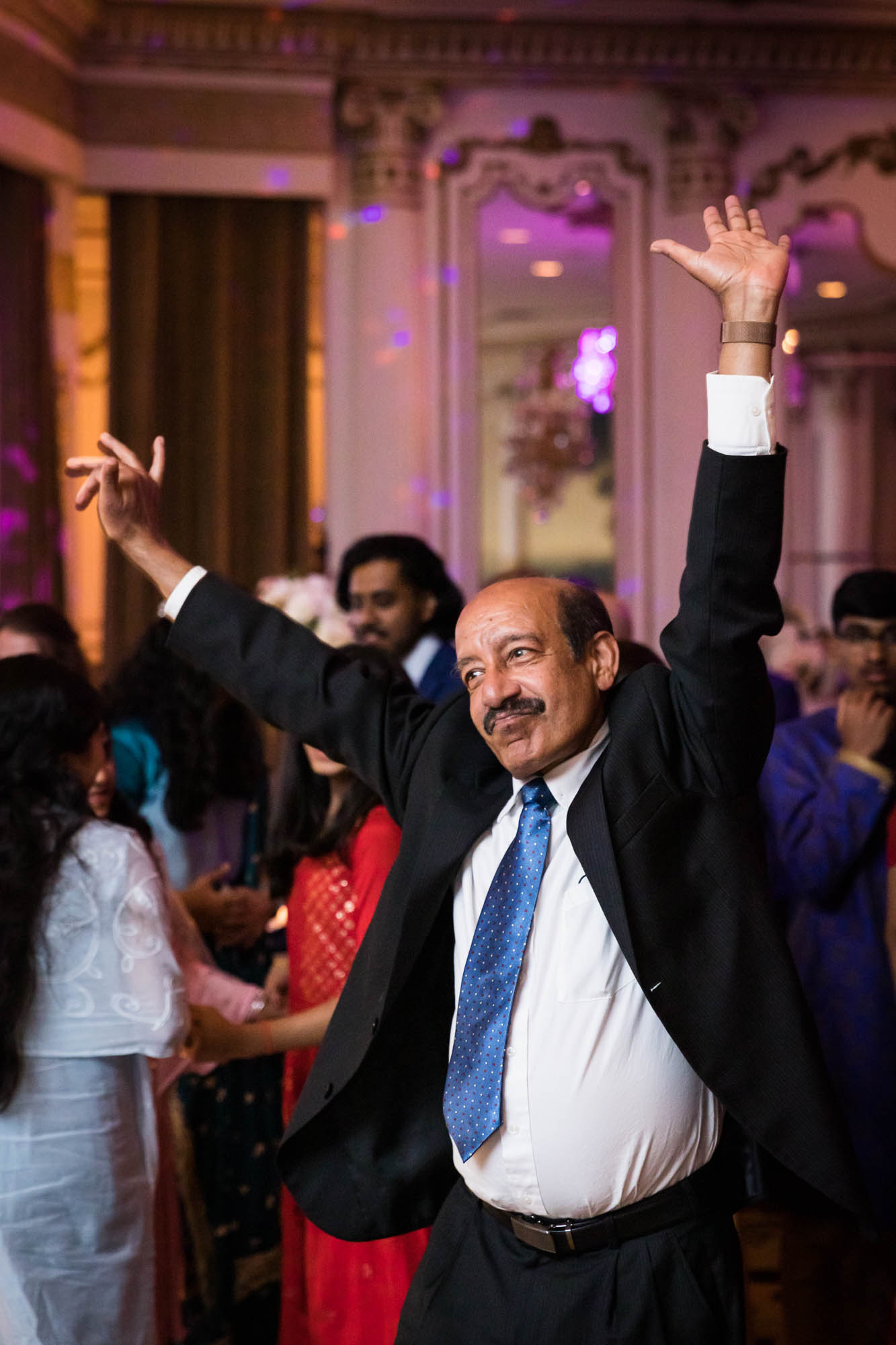 Bald Indian man wearing black suit and blue tie dancing with arms raised overhead for an article on Terrace on the Park wedding photo tips