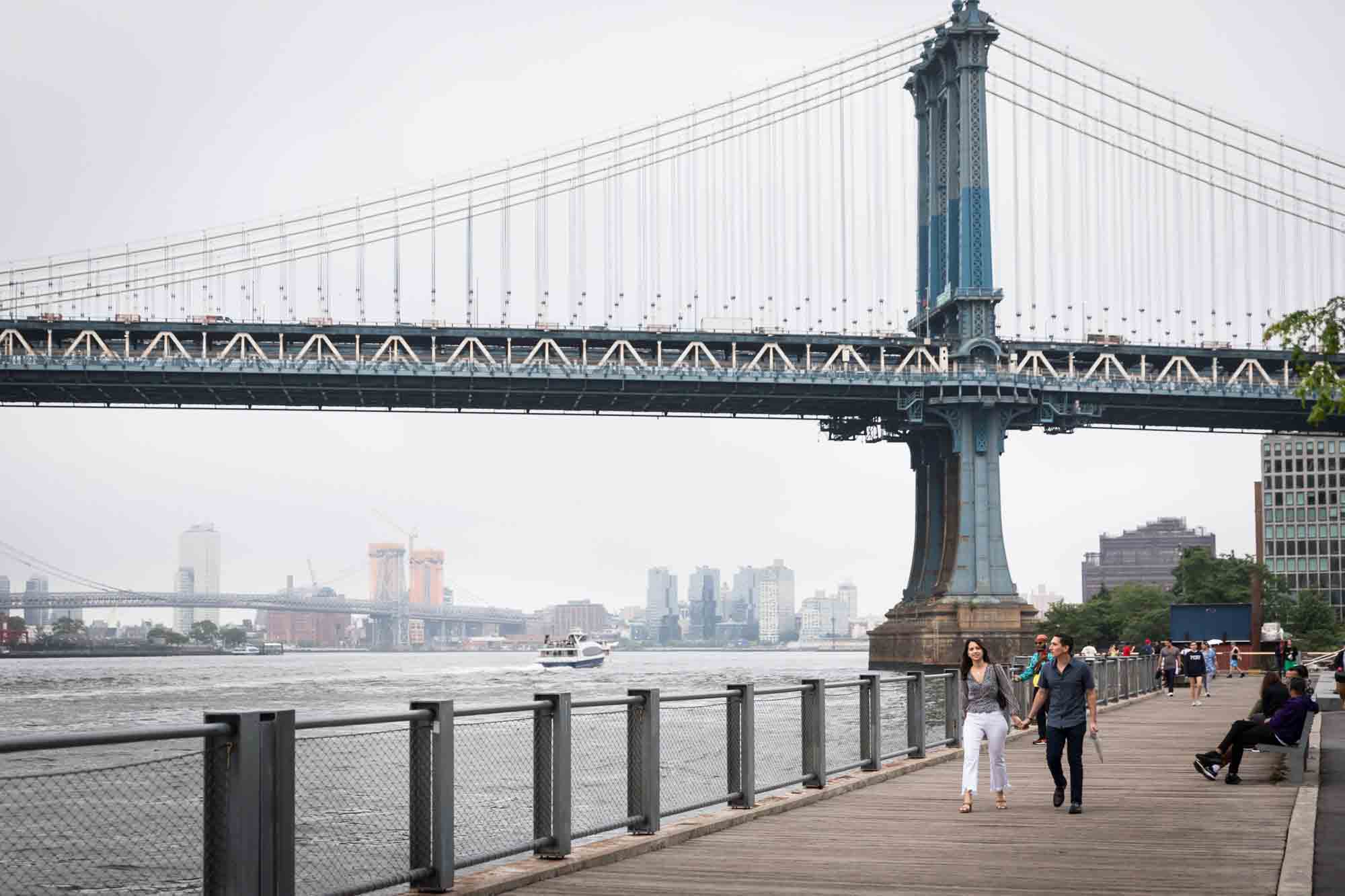 Couple walking along boardwalk in with Manhattan Bridge in background for an article on Brooklyn Bridge Park rainy day photo locations
