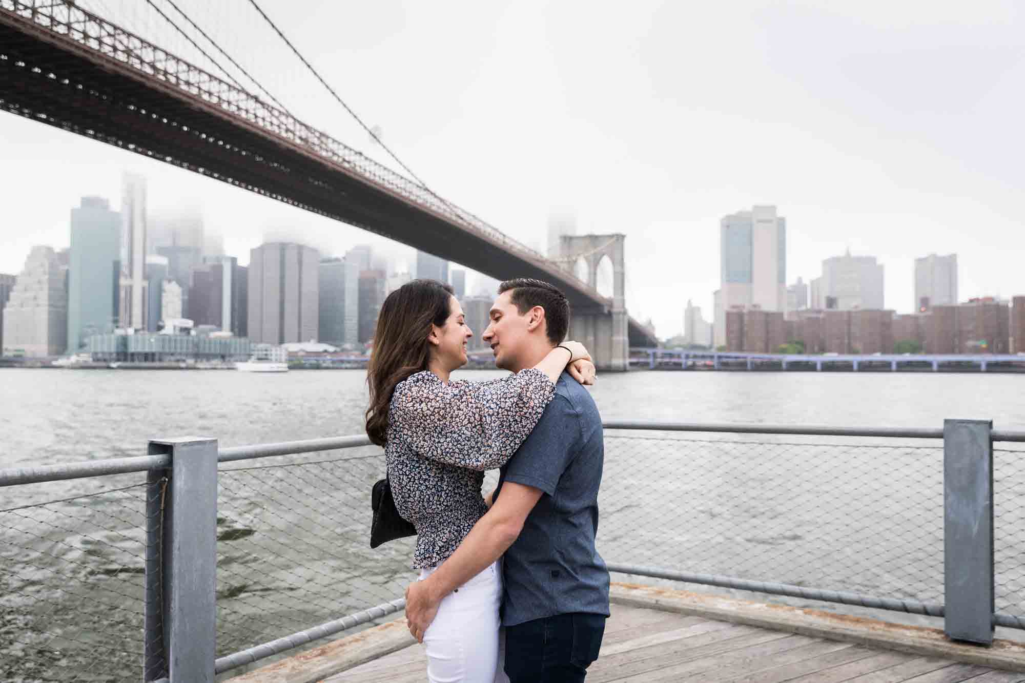 Couple hugging on waterfront with Brooklyn Bridge in background for an article on Brooklyn Bridge Park rainy day photo locations