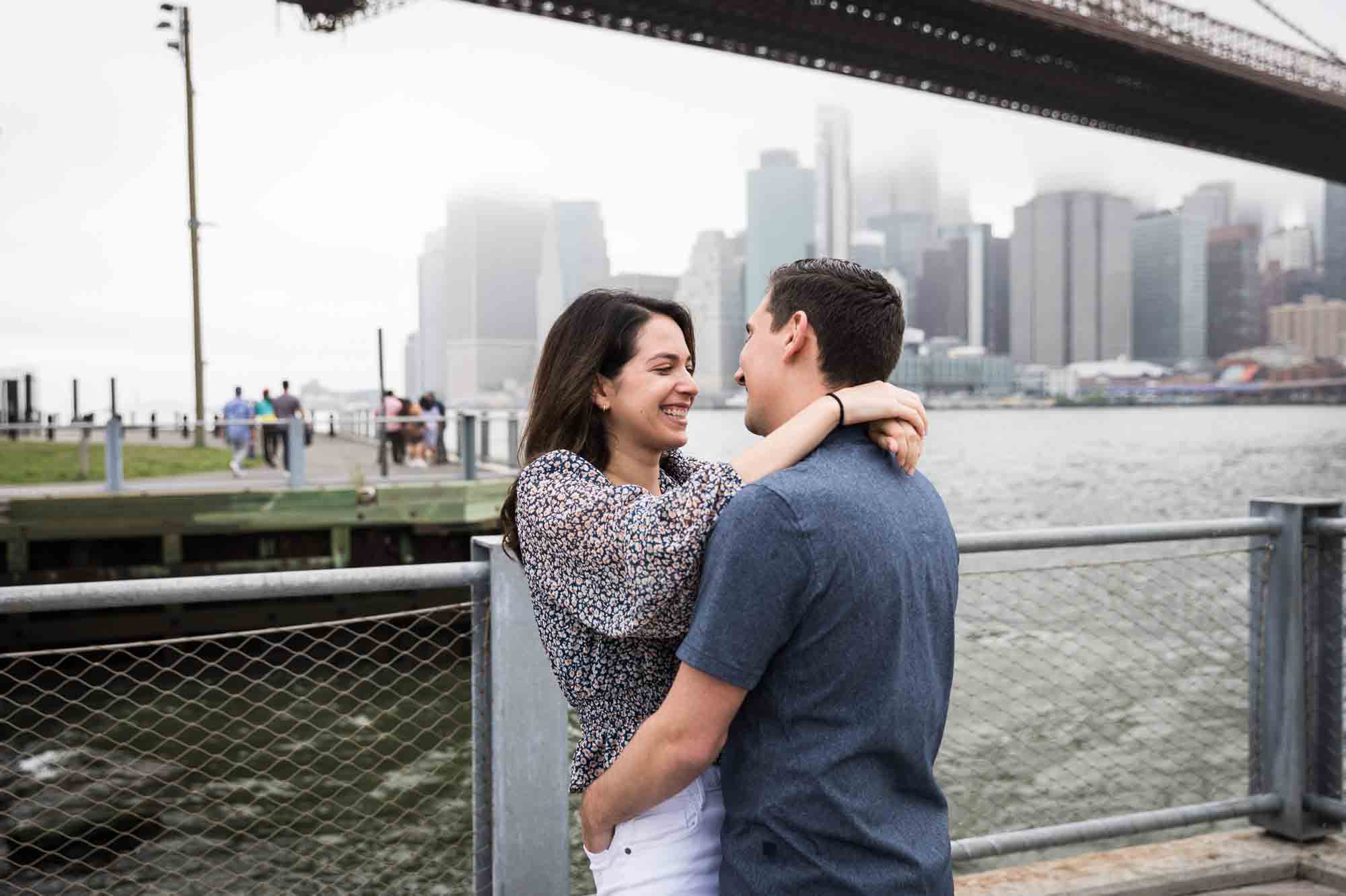Couple hugging on waterfront with Brooklyn Bridge in background for an article on Brooklyn Bridge Park rainy day photo locations