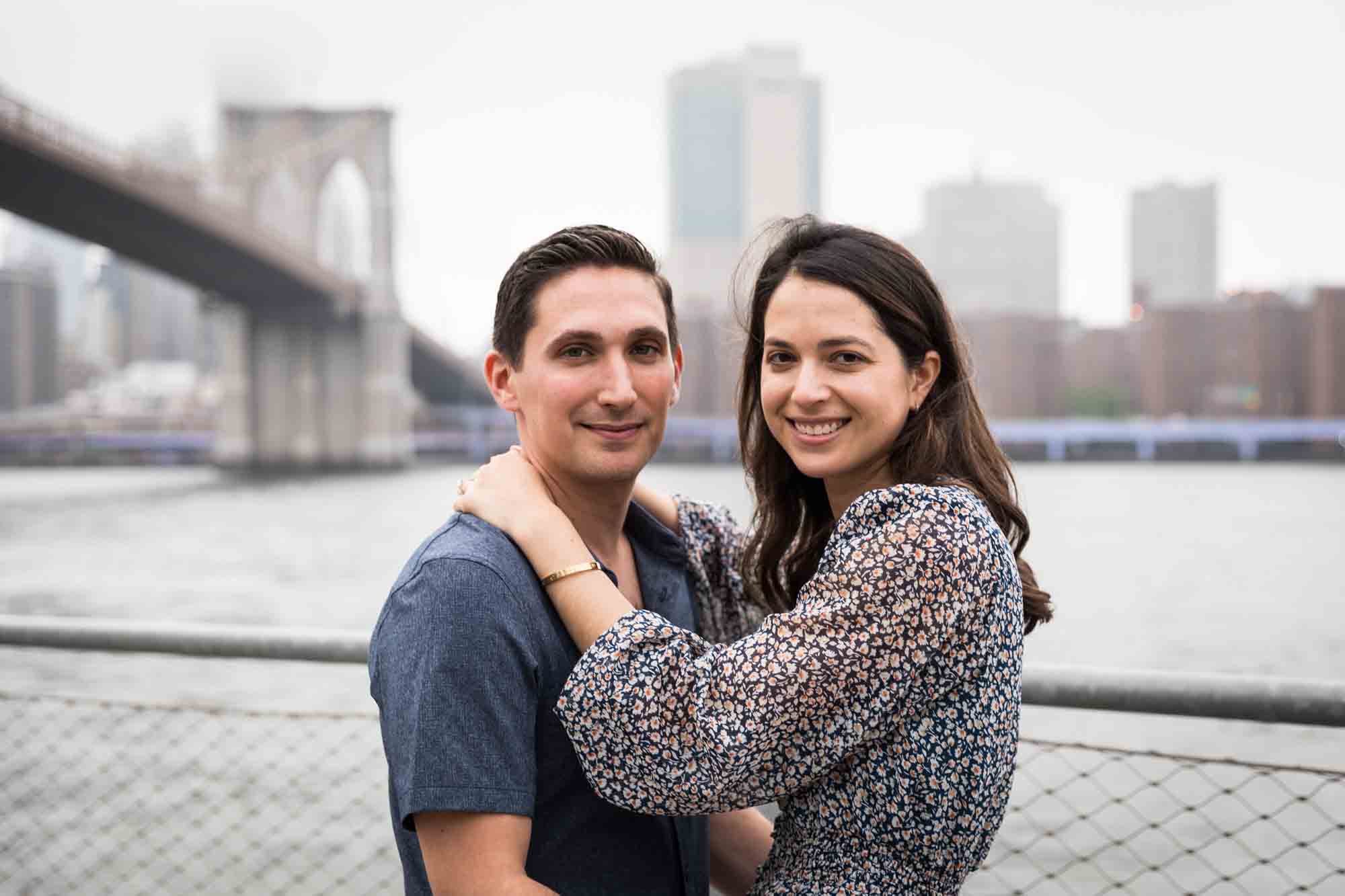 Couple hugging in front of waterfront and Brooklyn Bridge after a Brooklyn Bridge Park surprise proposal