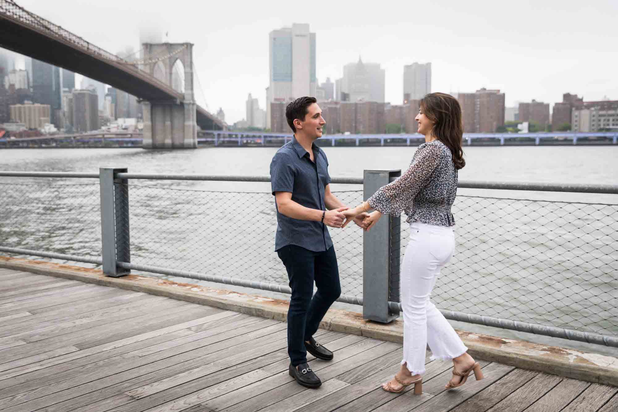 Couple dancing on boardwalk in front of waterfront and Brooklyn Bridge after a Brooklyn Bridge Park surprise proposal