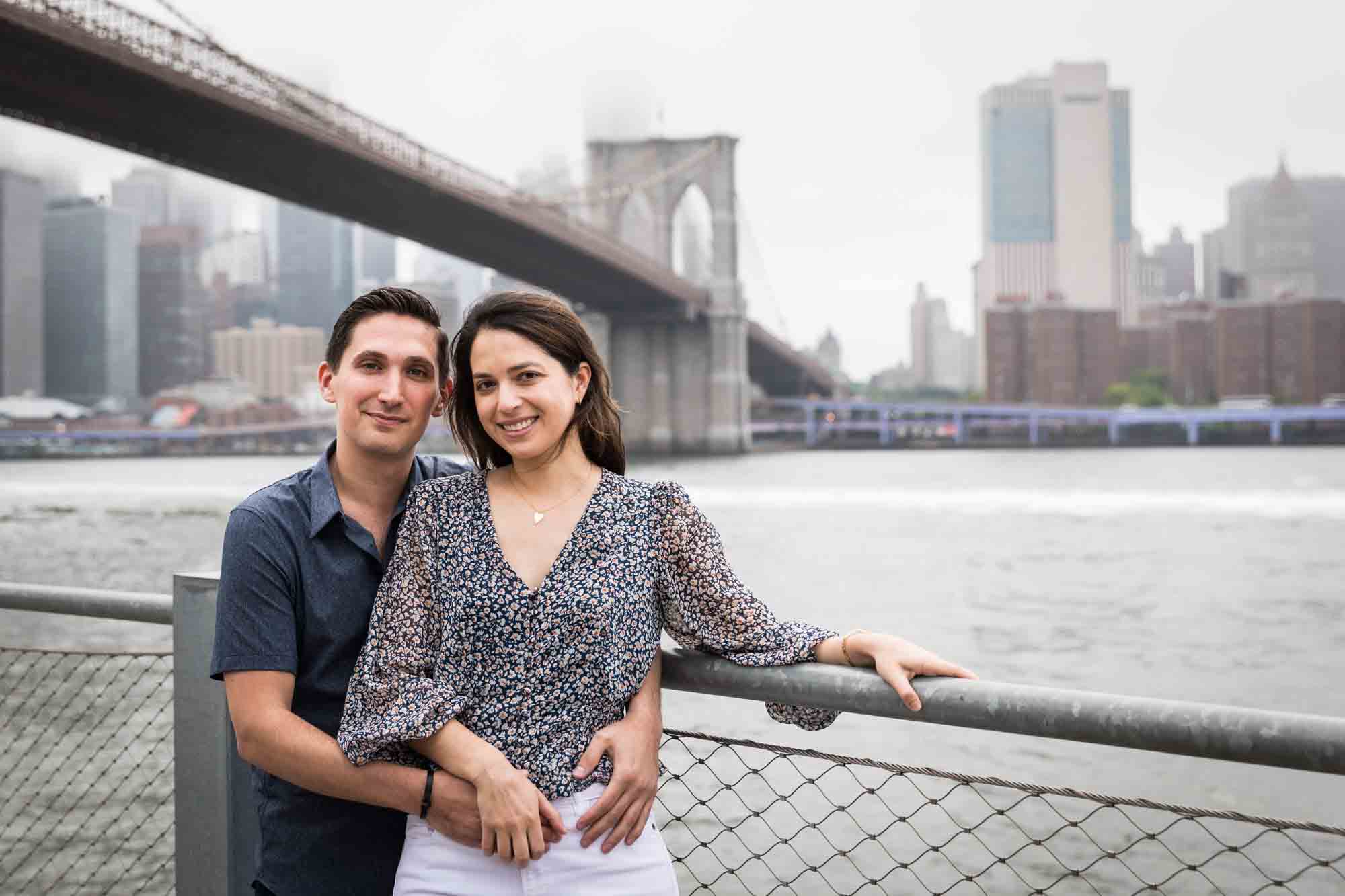 Couple standing in front of waterfront railing and Brooklyn Bridge after a Brooklyn Bridge Park surprise proposal
