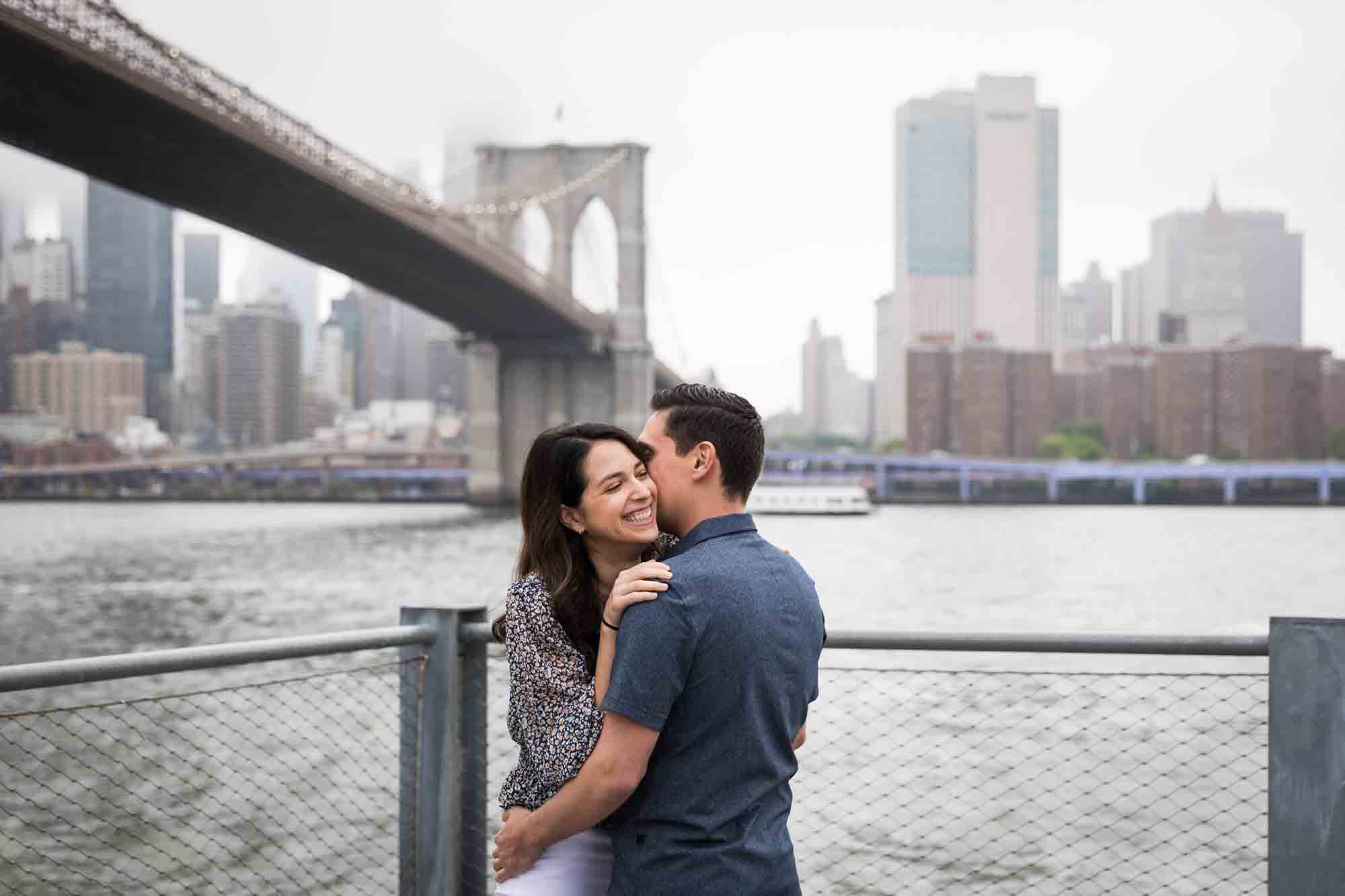 Couple hugging on waterfront with Brooklyn Bridge in background for an article on Brooklyn Bridge Park rainy day photo locations