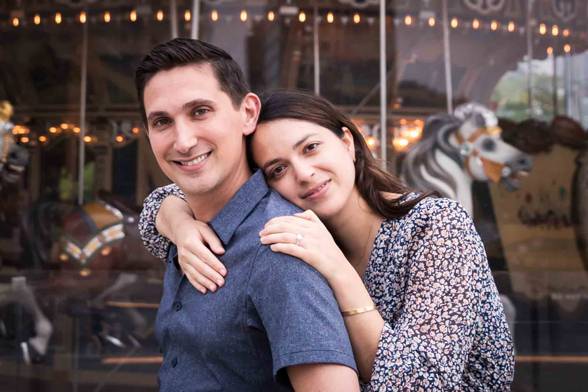 Woman leaning on man's back in front of Jane's Carousel after a Brooklyn Bridge Park surprise proposal