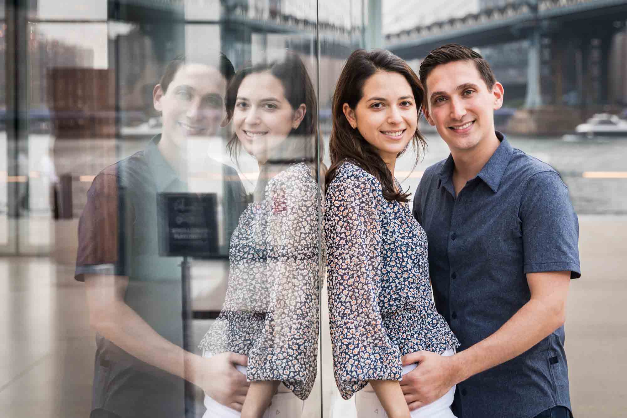 Couple leaning against glass wall with reflection after a Brooklyn Bridge Park surprise proposal