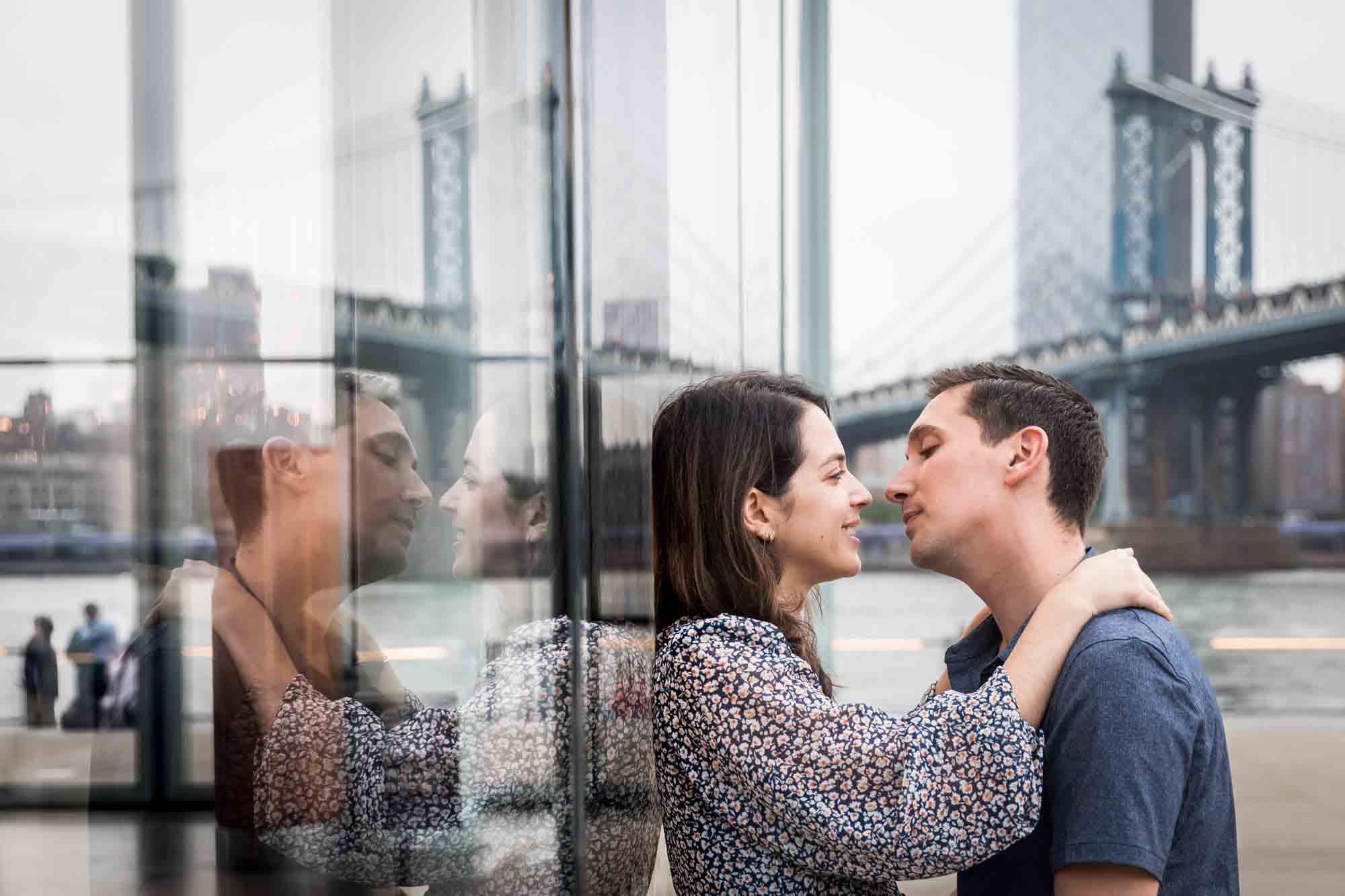 Couple about to kiss against glass wall with reflection and Manhattan Bridge in background after a Brooklyn Bridge Park surprise proposal