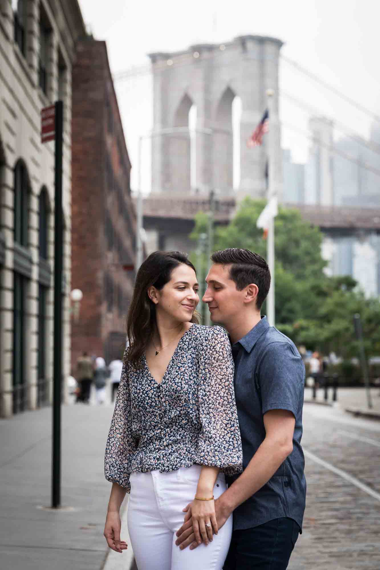 Couple hugging in middle of cobblestone street with Brooklyn Bridge in background after a Brooklyn Bridge Park surprise proposal