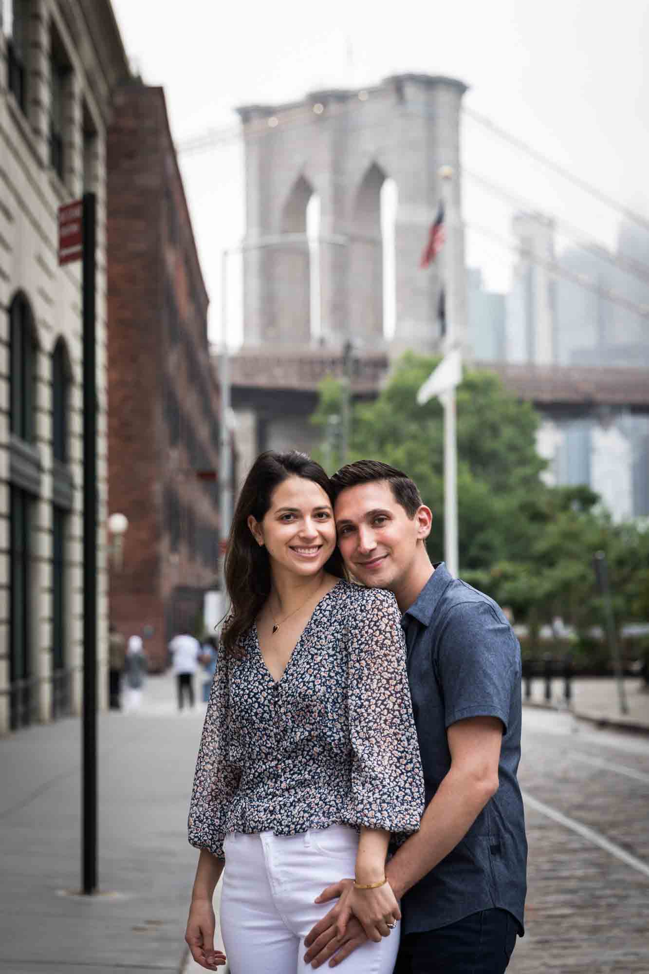Couple hugging in middle of cobblestone street with Brooklyn Bridge in background after a Brooklyn Bridge Park surprise proposal