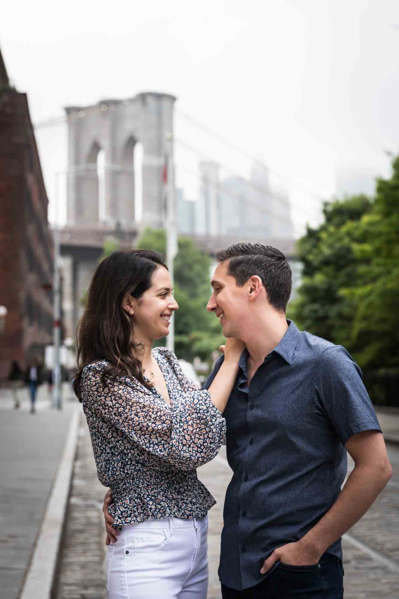 Couple hugging in middle of cobblestone street with Brooklyn Bridge in background after a Brooklyn Bridge Park surprise proposal