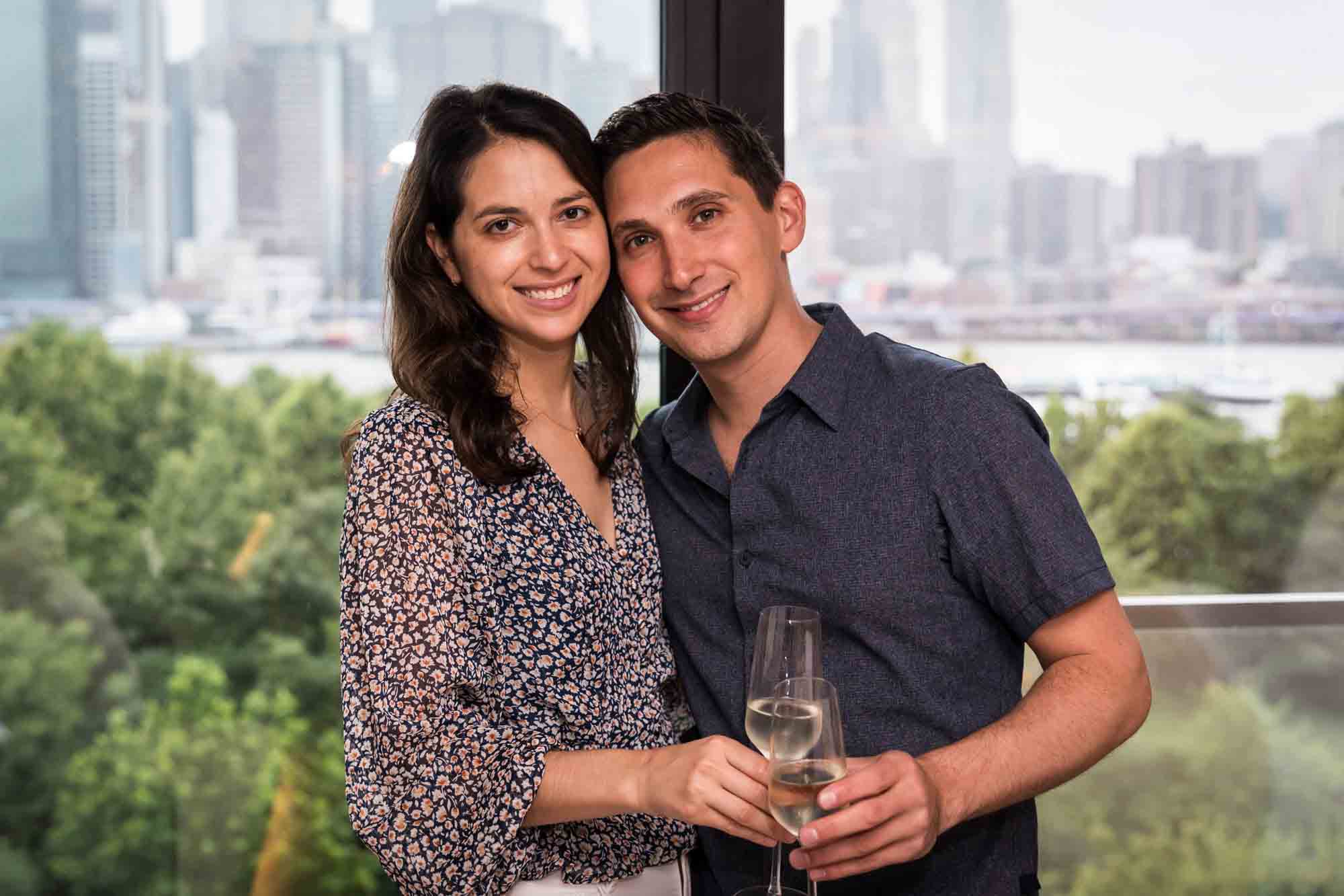 Couple standing together holding champagne glasses in front of window with NYC skyline in background