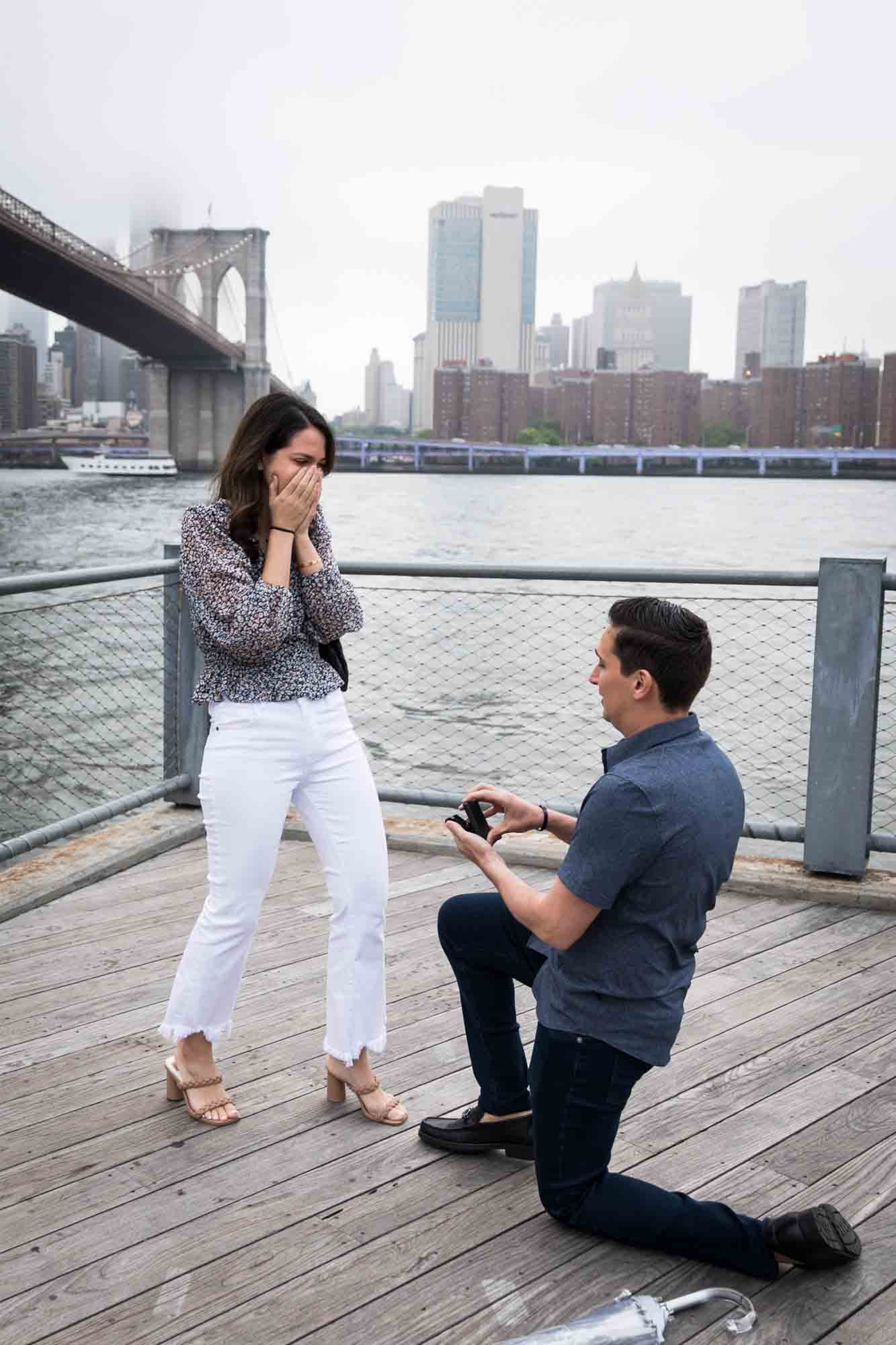 Man down on one knee with ring box in hand in front of woman with hands covering her face for an article on Brooklyn Bridge Park rainy day photo locations