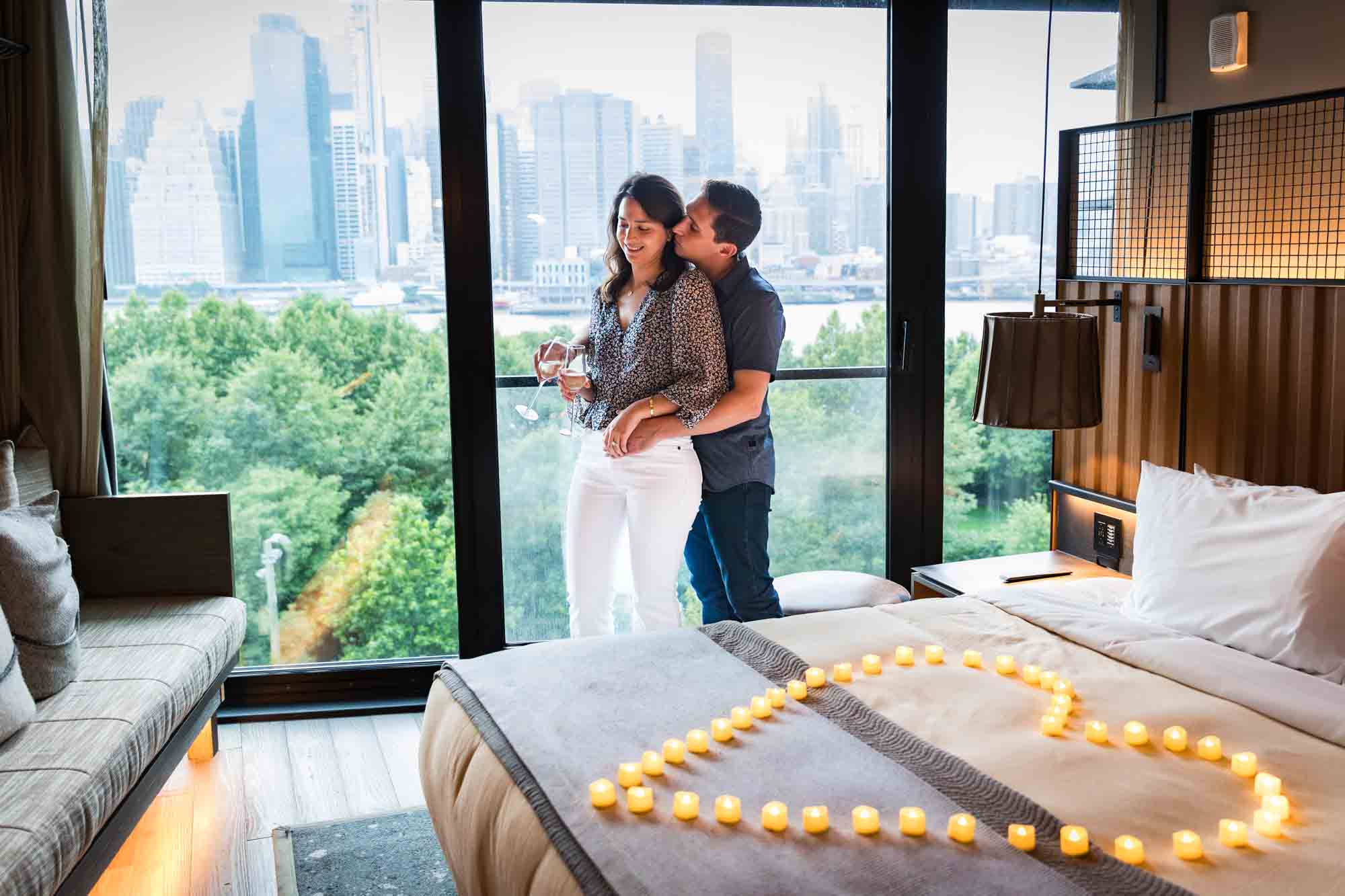 Couple standing together holding champagne glasses in front of hotel room window with NYC skyline in background