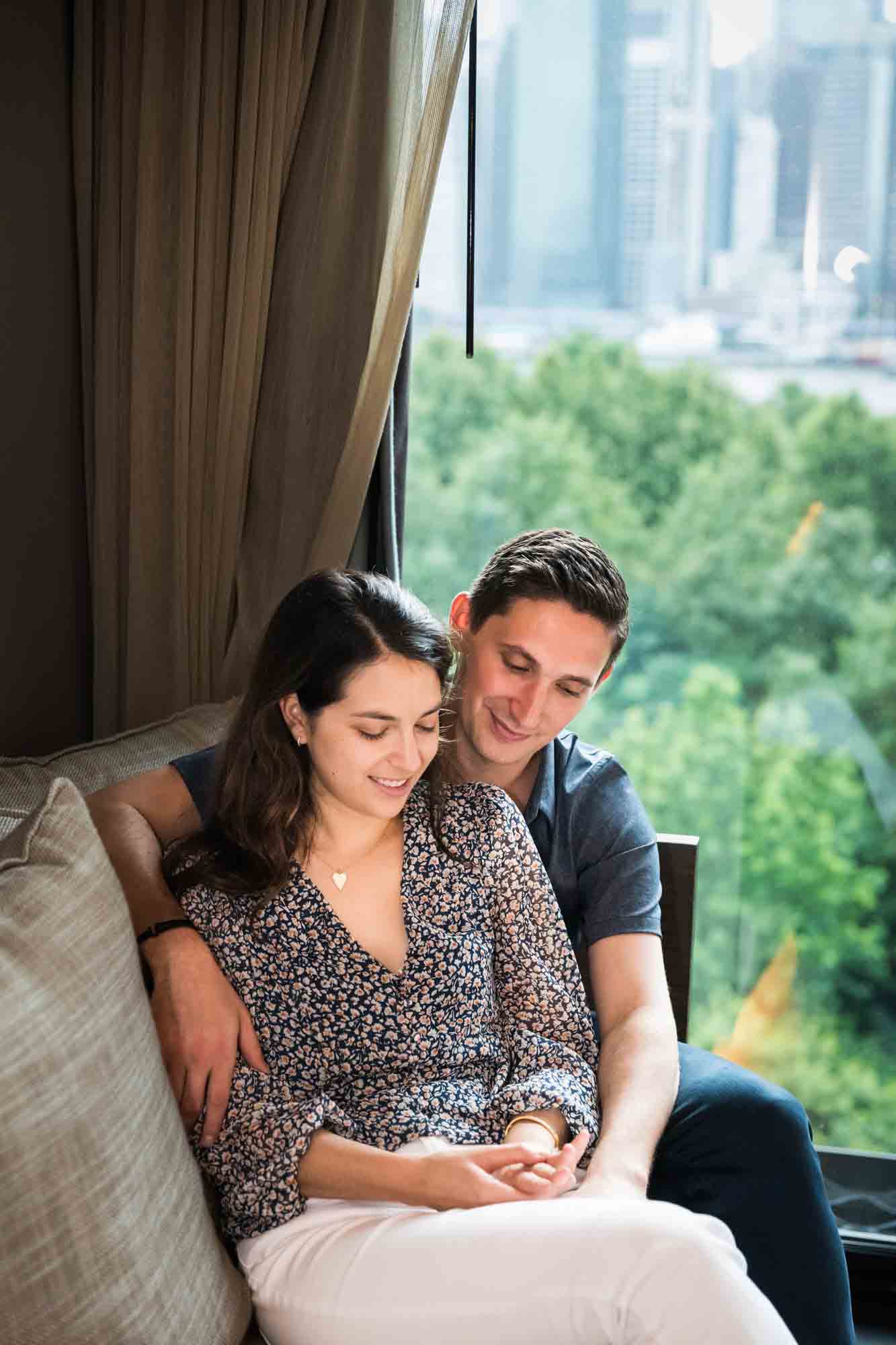 Couple sitting together on couch looking at engagement ring on woman's hand in front of large window at 1 Brooklyn Bridge Hotel