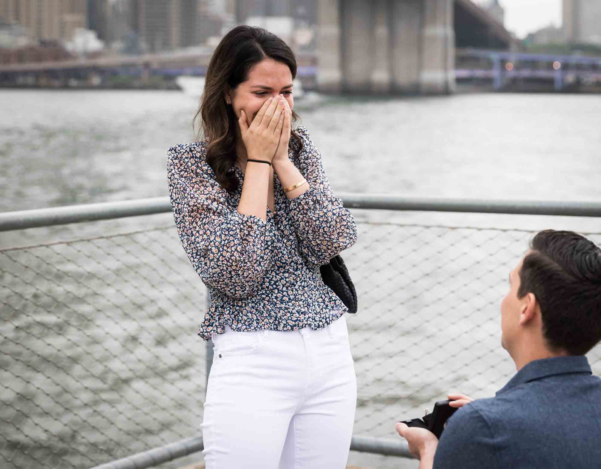 Woman with hands covering her face in shock in front of man holding engagement ring box for an article on Brooklyn Bridge Park rainy day photo locations