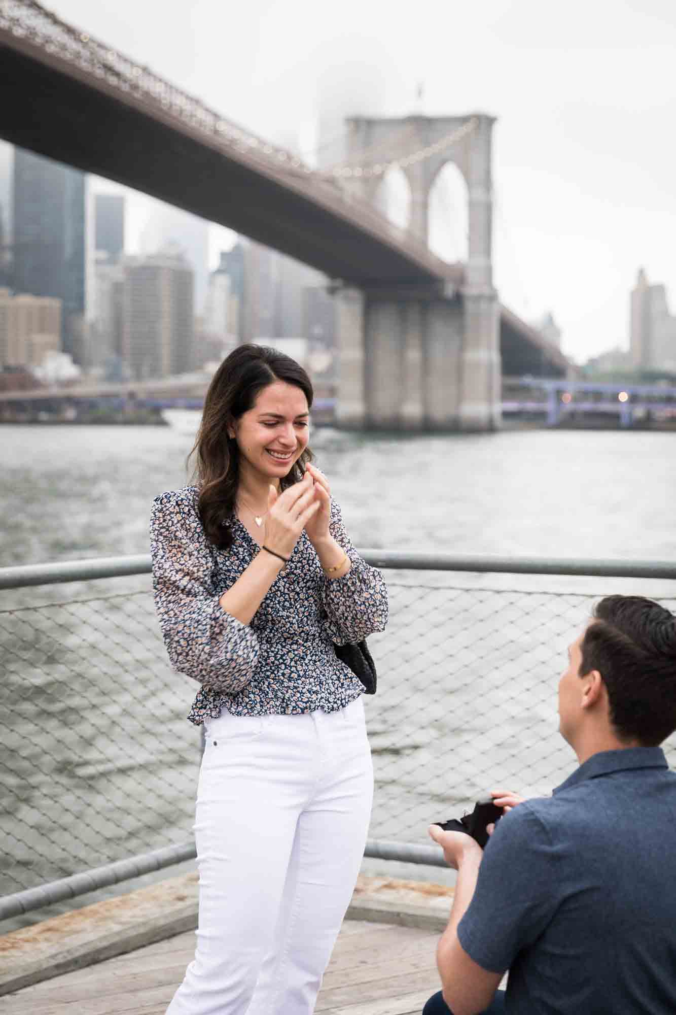 Woman looking shocked standing in front of man holding engagement ring box for an article on Brooklyn Bridge Park rainy day photo locations
