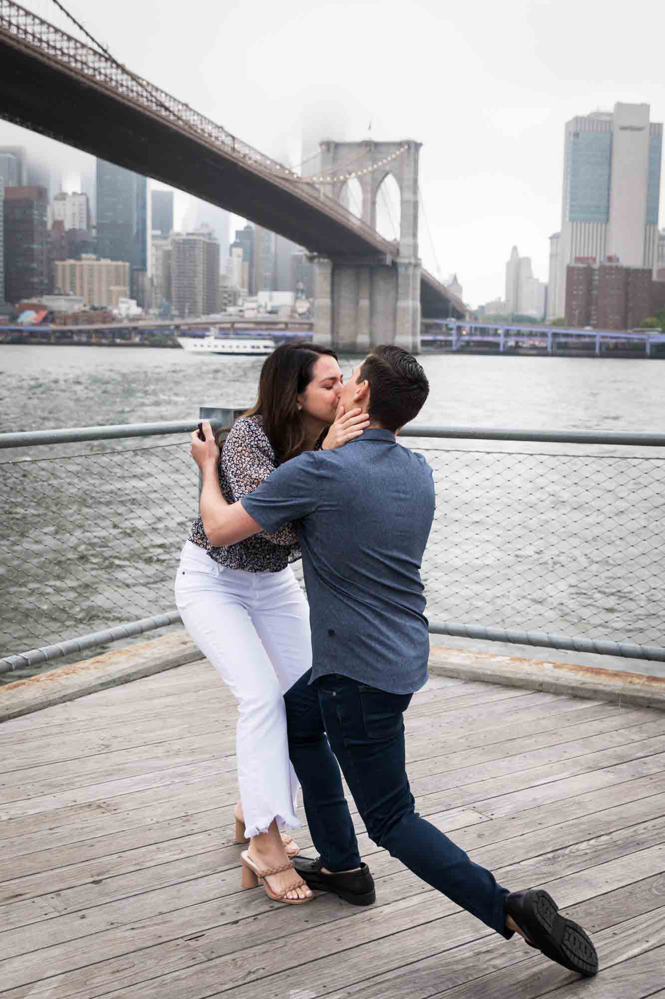 Woman kissing man on boardwalk in front of Brooklyn Bridge for an article on Brooklyn Bridge Park rainy day photo locations