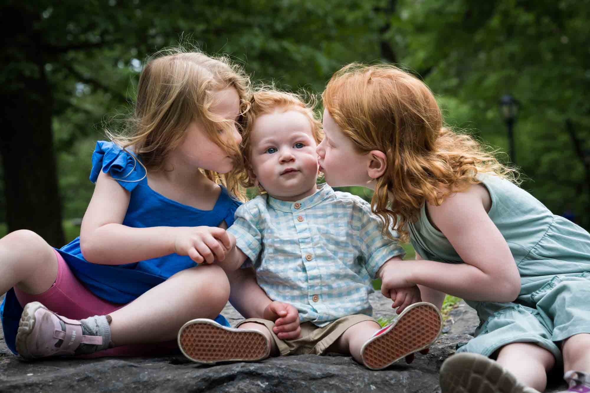 Two little girls kissing red-haired baby boy sitting on rock during a Central Park family portrait session