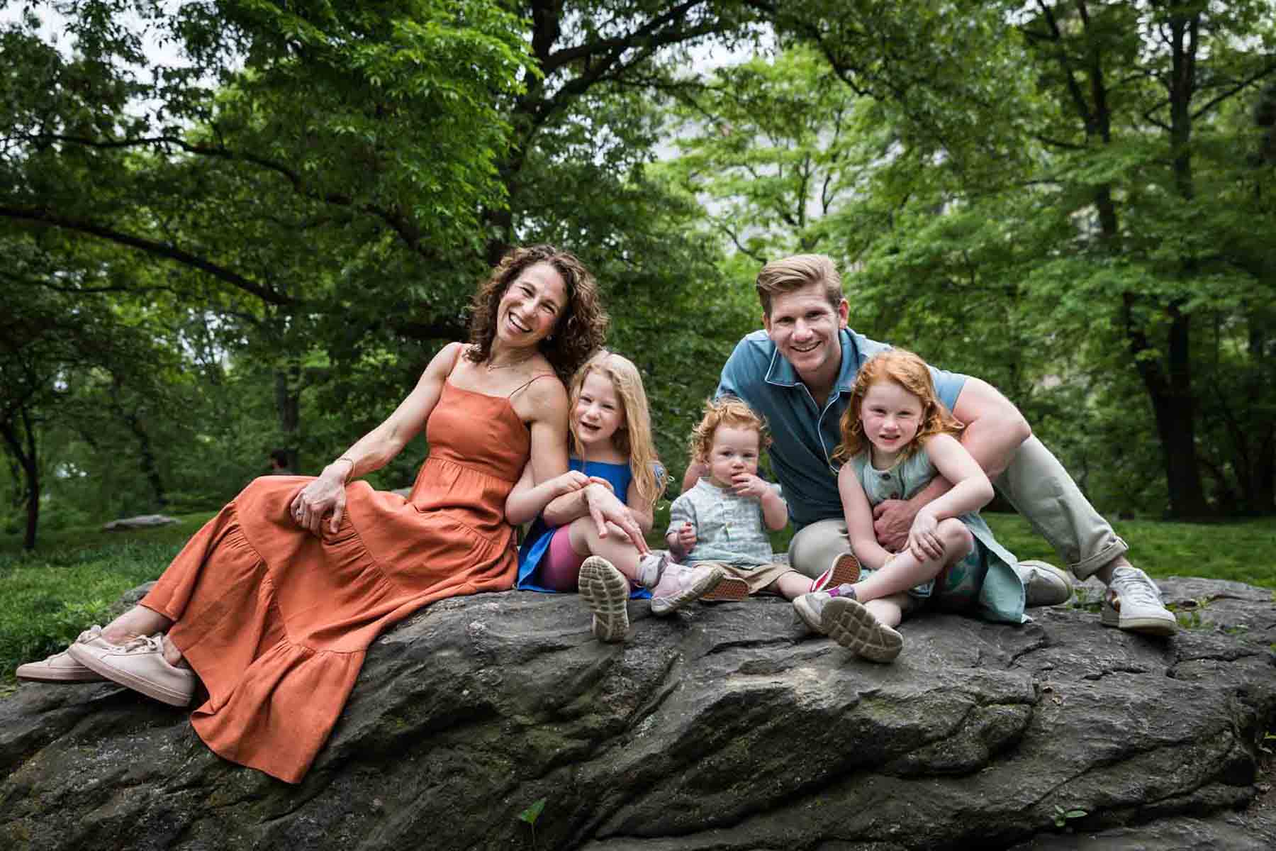 Mother wearing orange dress and father with three children sitting on rock during a Central Park family portrait session