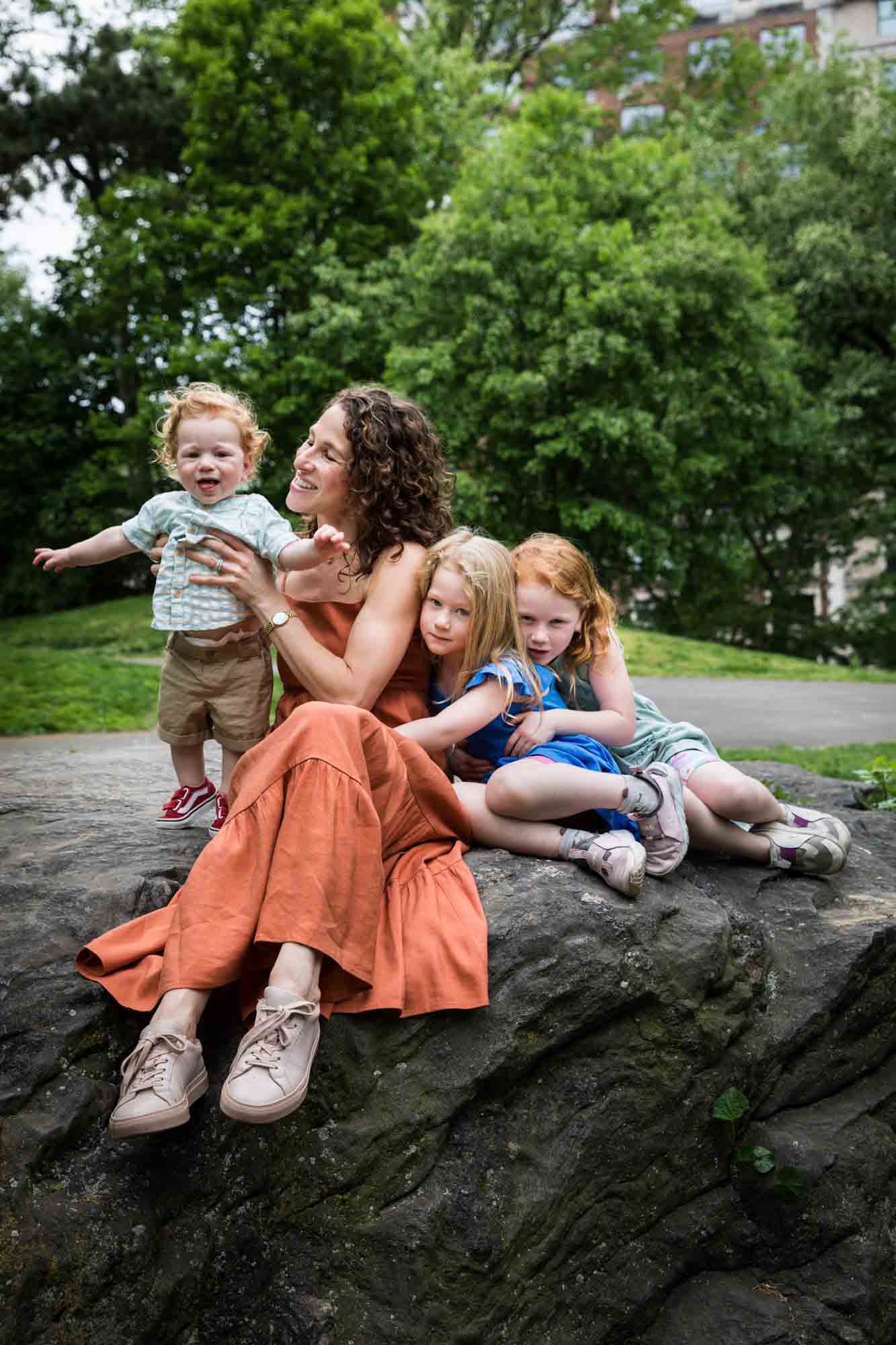 Mother wearing orange dress with three children sitting on rock during a Central Park family portrait session