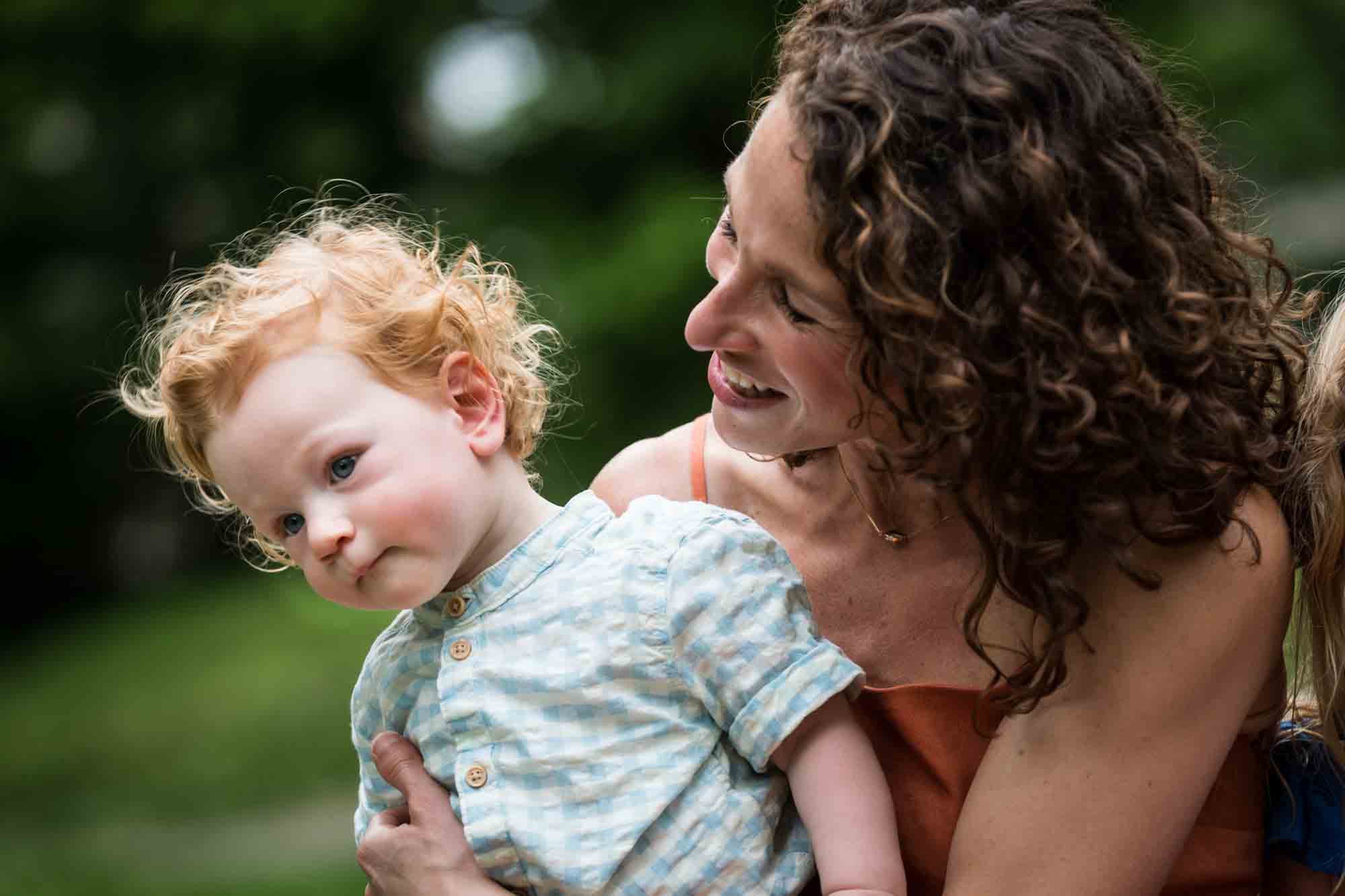 Mother wearing orange dress holding red-haired baby boy in front of trees during a Central Park family portrait session