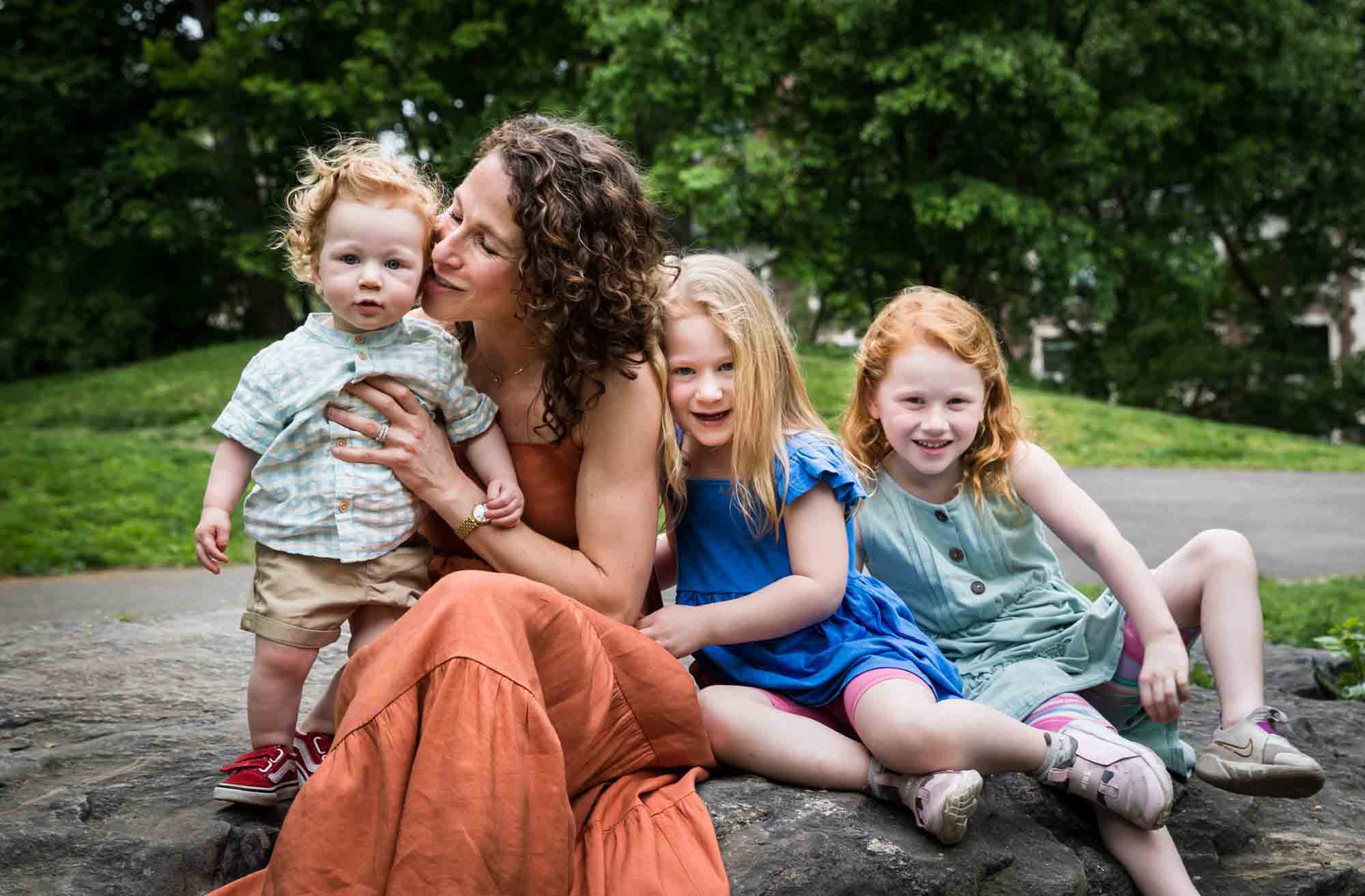 Mother wearing orange dress with three children sitting on rock during a Central Park family portrait session