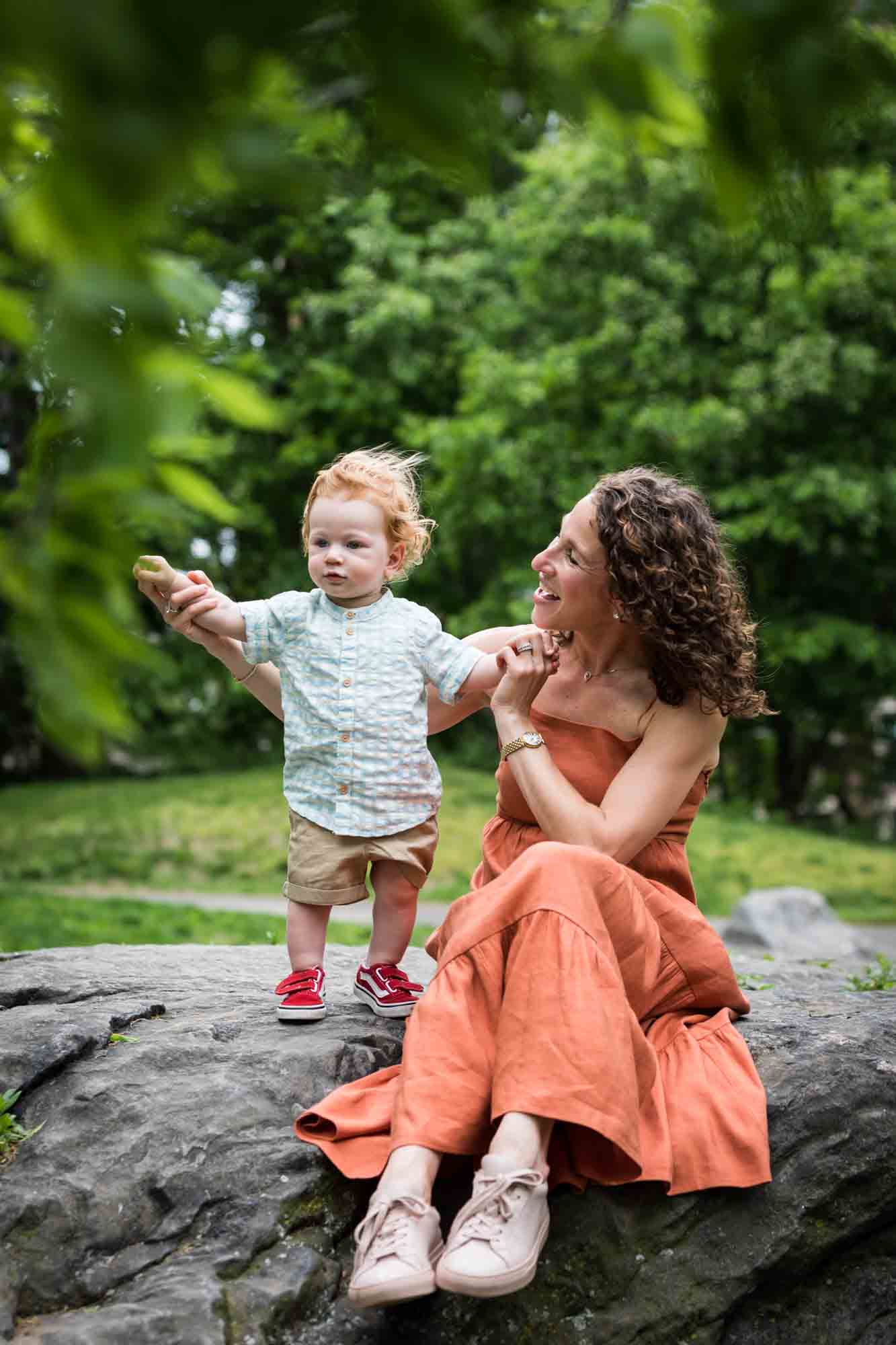 Mother wearing orange dress holding red-haired baby boy on rock during a Central Park family portrait session