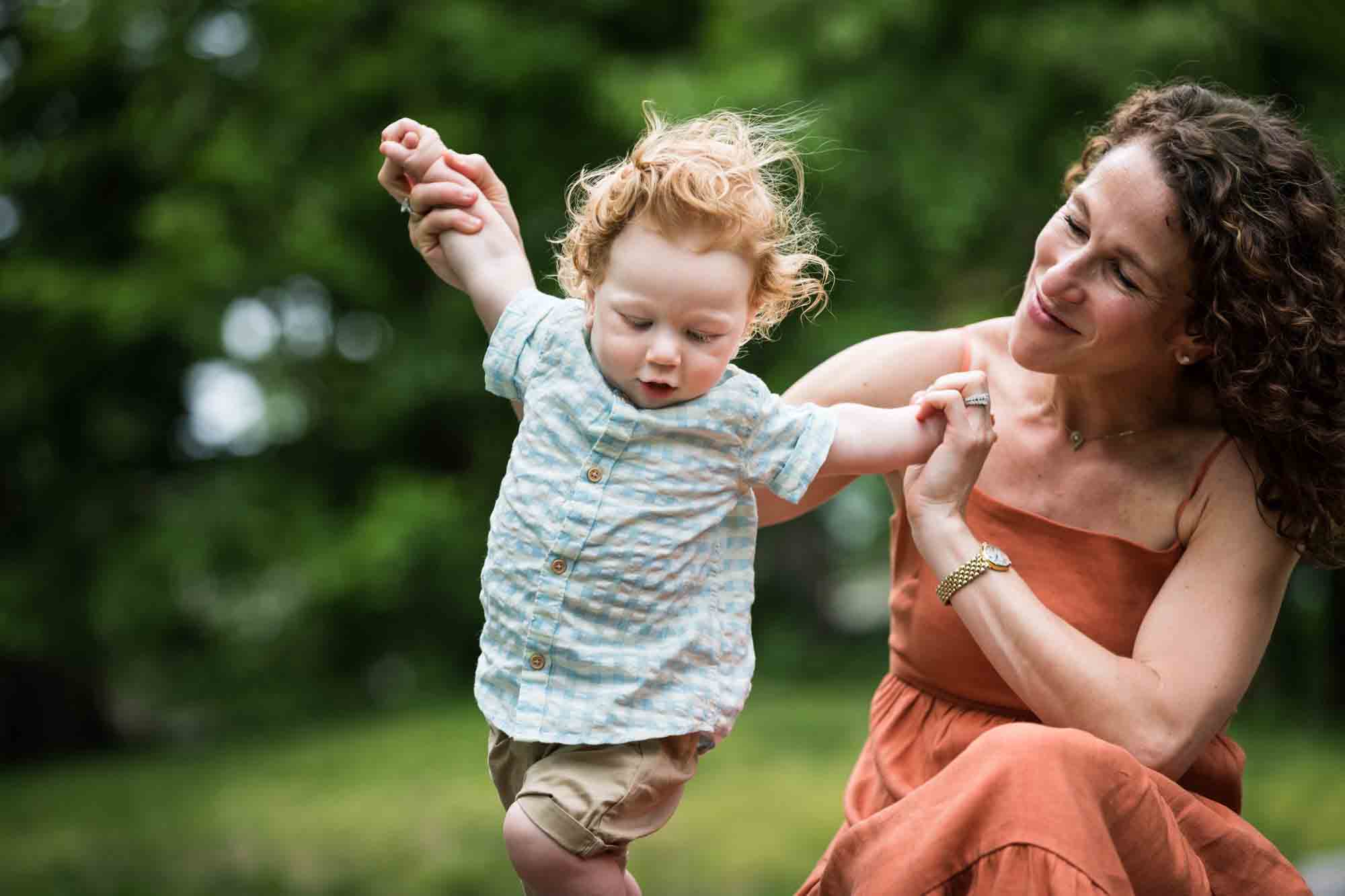 Mother wearing orange dress holding red-haired baby boy in front of trees during a Central Park family portrait session