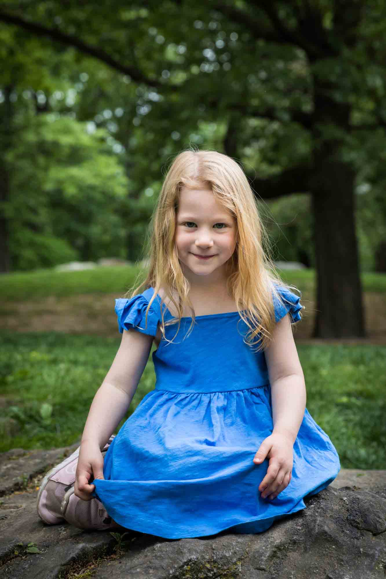 Blonde-haired little girl wearing blue dress sitting on rock during a Central Park family portrait session