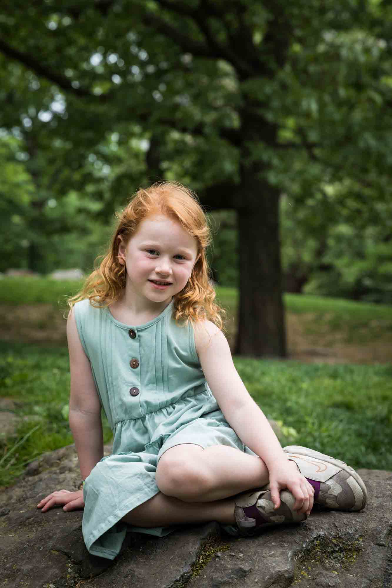 Red-haired little girl wearing green dress sitting on rock during a Central Park family portrait session