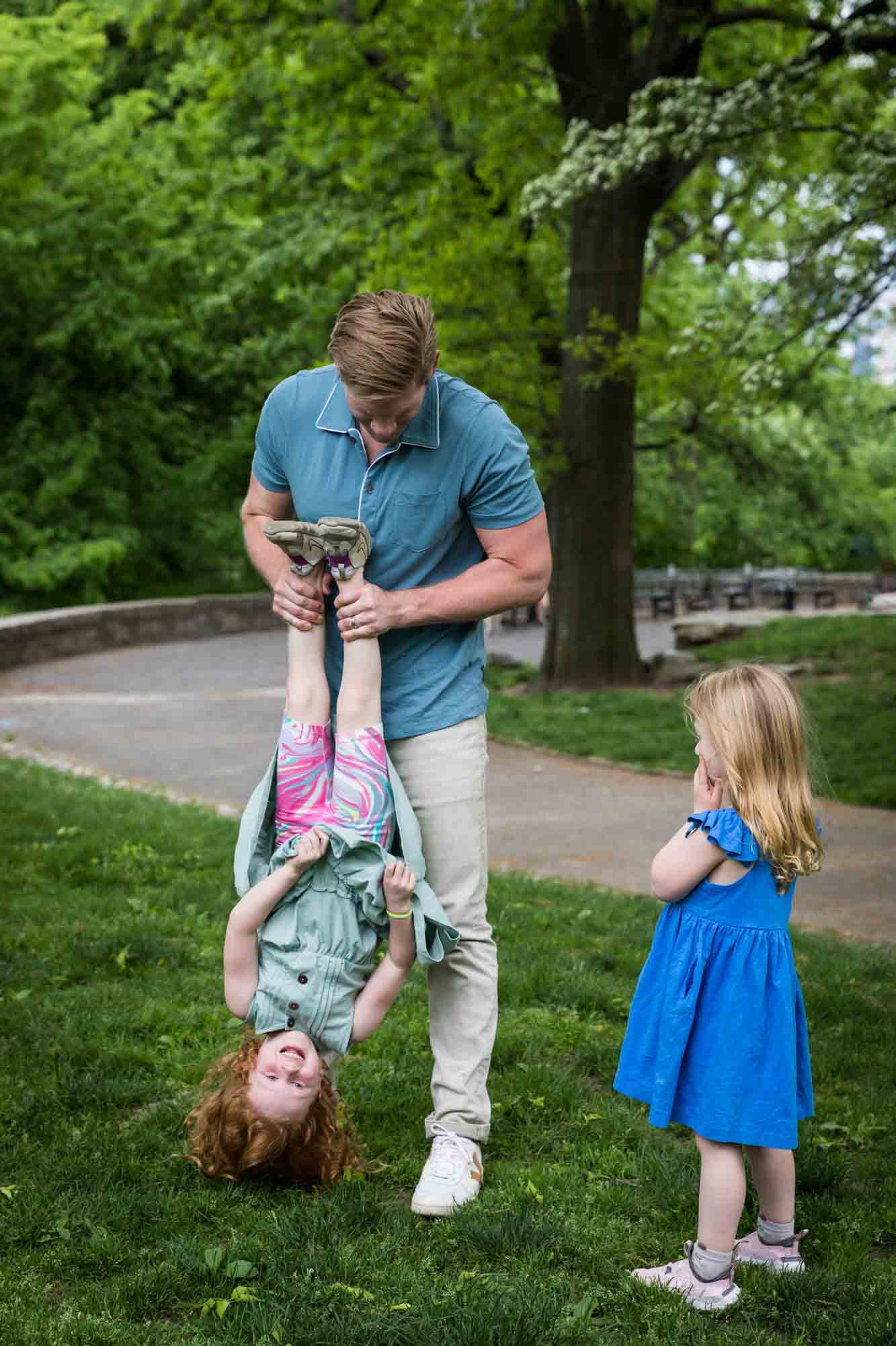 Father holding daughter upside down in the grass with girl wearing blue dress watching