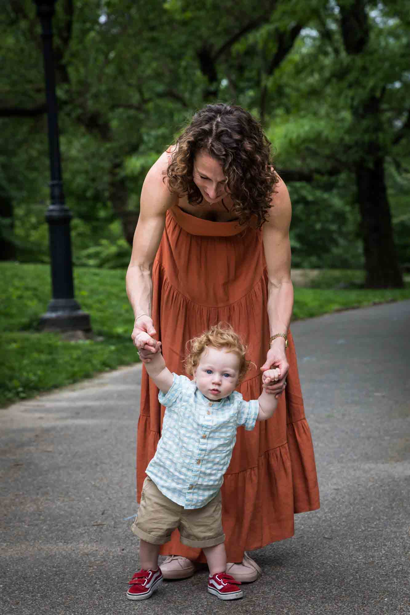 Mother wearing orange dress helping red-haired baby boy walk down a pathway during a Central Park family portrait session