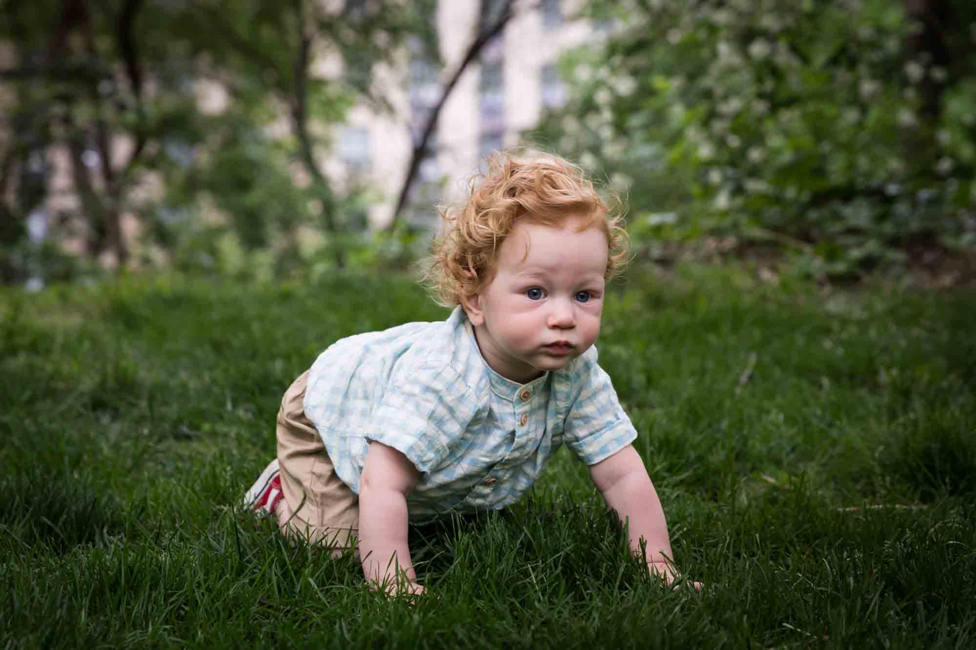 Red-haired baby boy wearing blue and white checked shirt crawling in the grass during a Central Park family portrait session