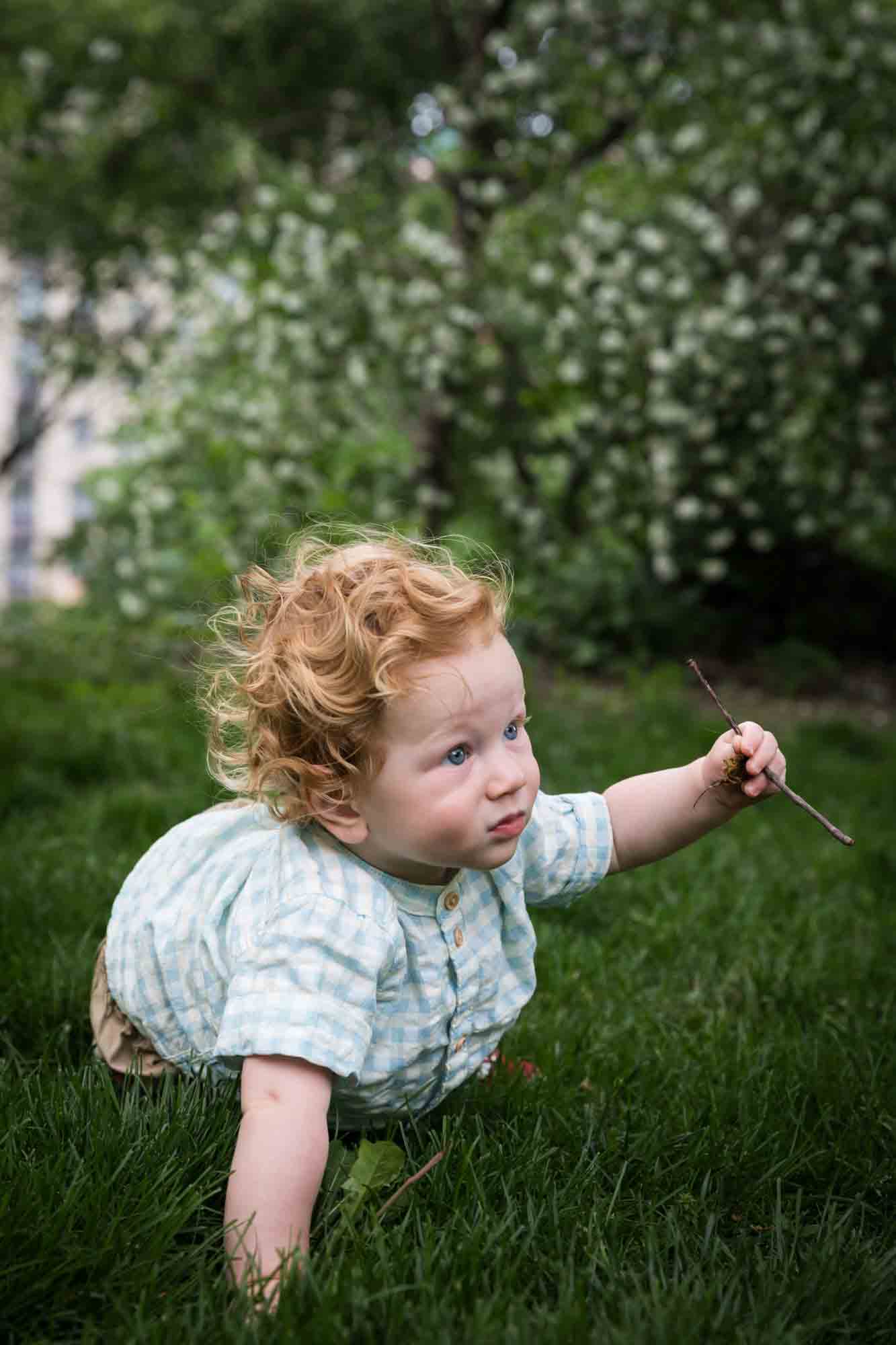 Red-haired baby boy wearing blue and white checked shirt sitting in the grass holding a stick during a Central Park family portrait session